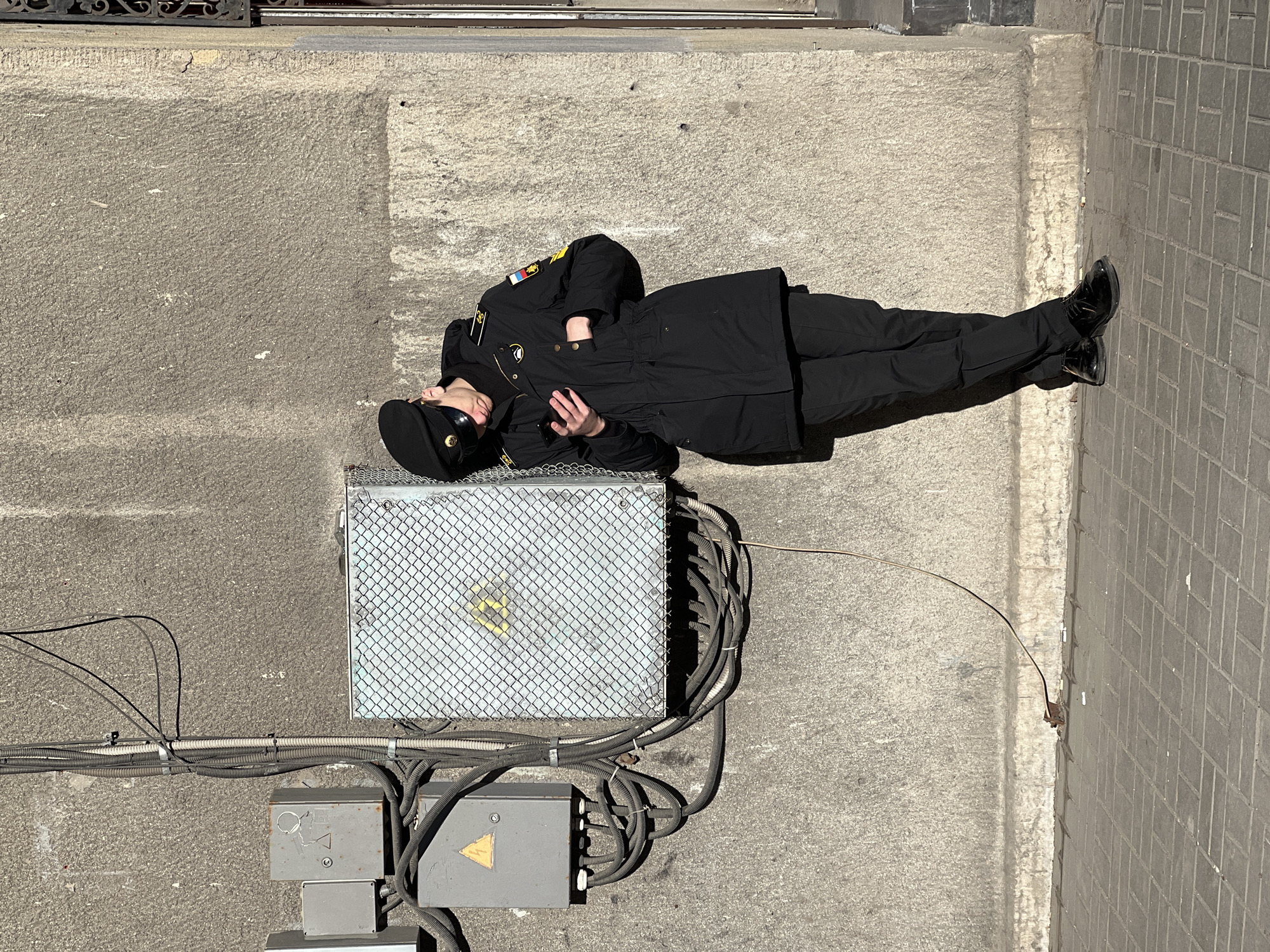 A person in a dark uniform is lying on a gray metal box with electrical conduits attached, set on a concrete surface. The person wears a black uniform with shoulder patches of an unknown design, a black cap with a gold emblem, and shiny black shoes. The person's head rests on the metal box, with one hand in their pocket and the other resting near their chest. The concrete surface is next to a tiled walkway. The location is Saint Petersburg, Russia, as indicated by the Russian flag on the shoulder patch.