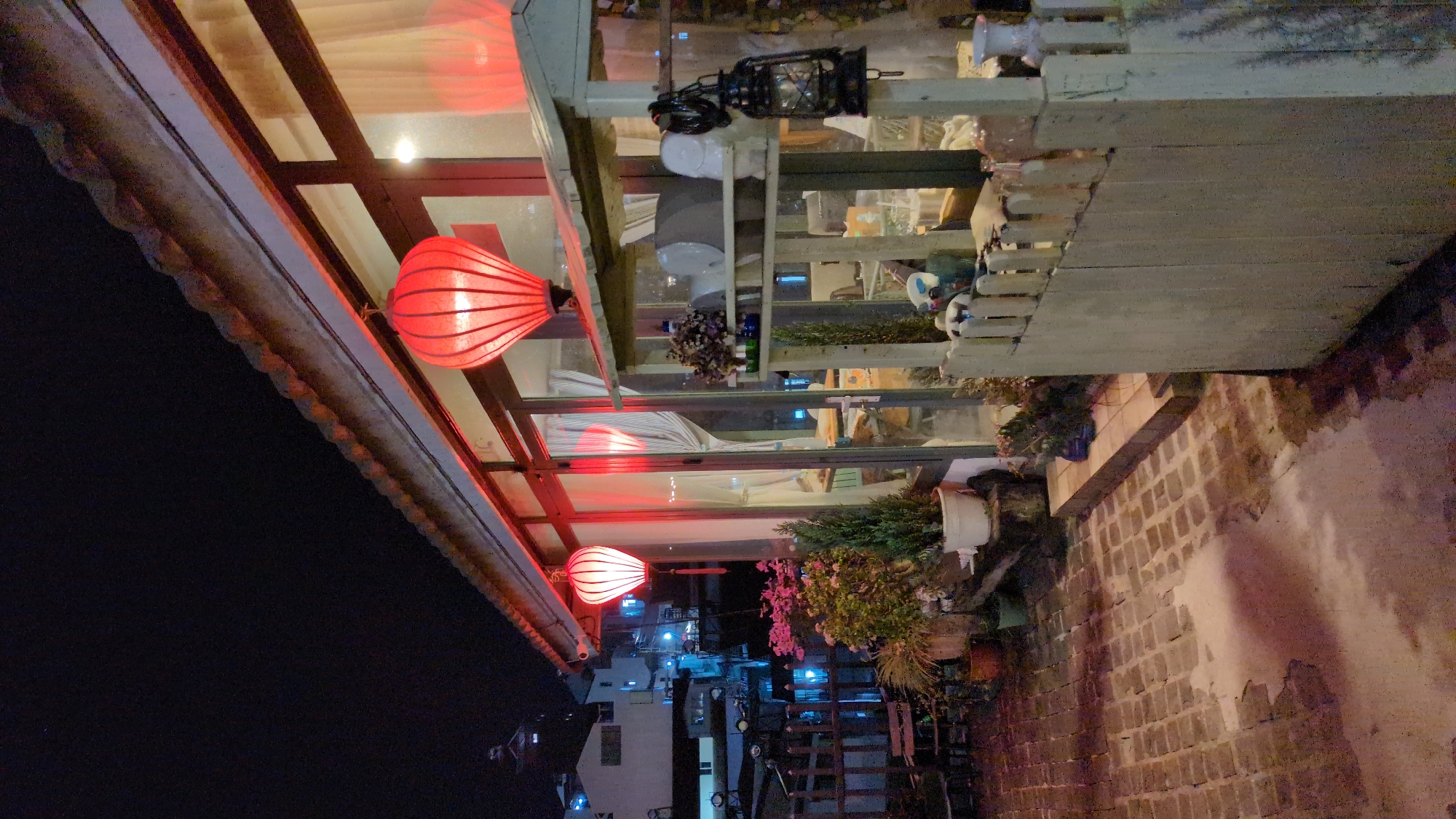 The image depicts an outdoor scene at night, likely in Da Lat, Vietnam, based on the architecture and lanterns. Three illuminated red lanterns are hung along the eaves of a building with a glass-enclosed space. The glass enclosure contains assorted items, including plants, bottles, and decorative objects. Outside, a brick path or courtyard is visible, with more potted plants and trellises along the edge. In the background, the city lights are visible, suggesting an urban setting.
