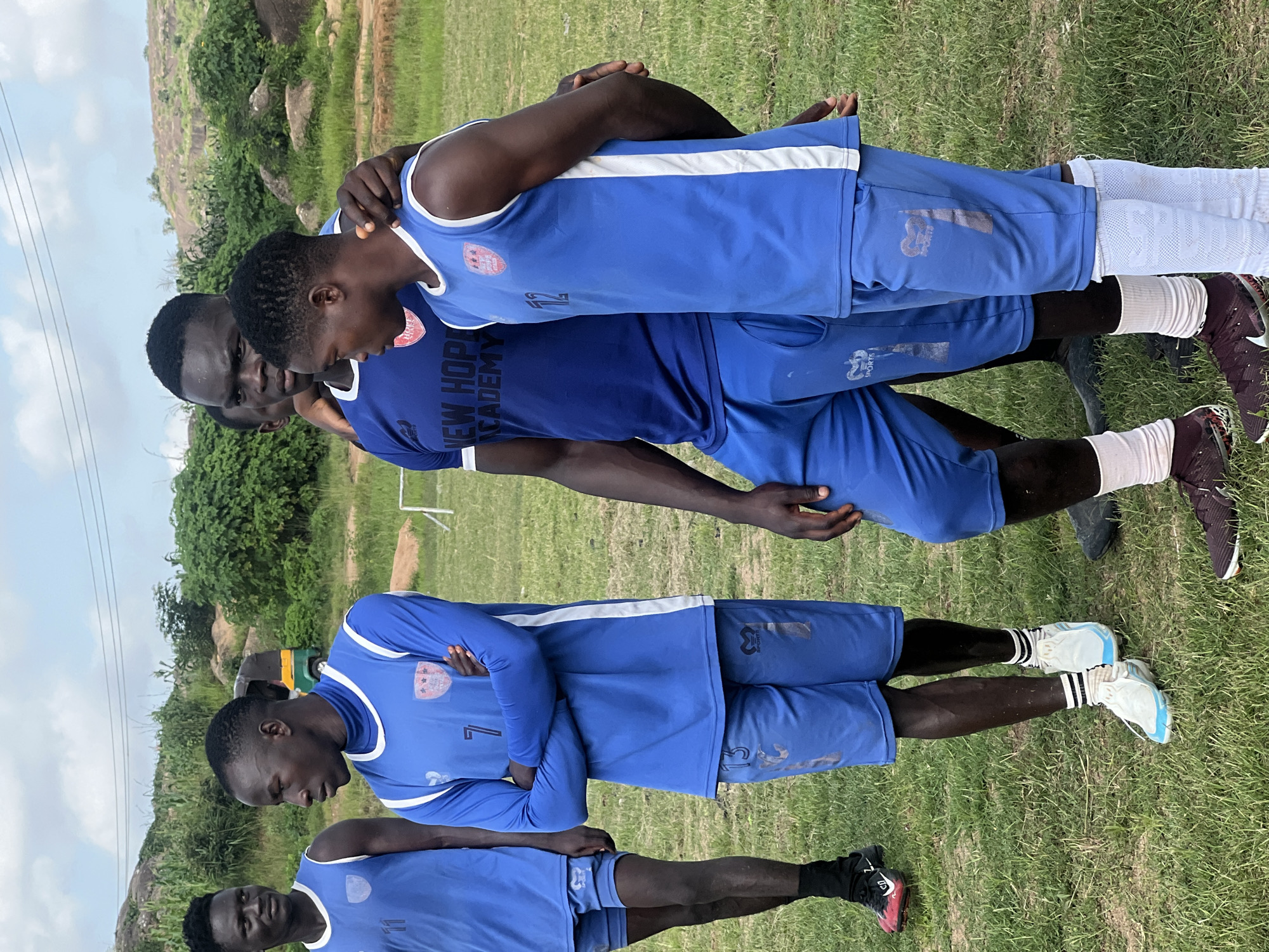 This photograph captures a group of young men, appearing to be a soccer team, gathered on a grassy field in Jos, Nigeria. The men are wearing blue athletic uniforms with white accents, and some have numbers visible on their jerseys and shorts. One individual in the center has his arm around another. The background shows a slightly hilly landscape with scattered green vegetation and a cloudy sky. A yellow and green autogyro is visible in the middle distance. The overall setting suggests an outdoor sports training or practice session.