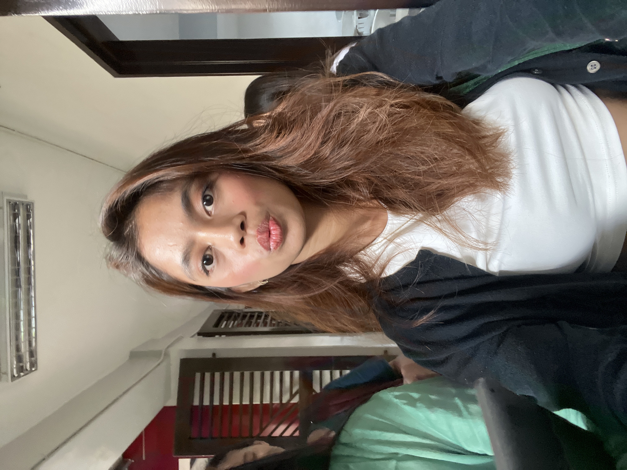 The image shows a selfie of a young woman with medium-length, light brown hair. She has tan skin, dark eyes, and is pursing her lips slightly. She is wearing a white t-shirt and a black jacket or cardigan. Part of a second person is visible in the lower portion of the frame. The background suggests an interior space, possibly a home, with a window or door featuring horizontal slats. The location is inferred to be Davao City, Philippines.