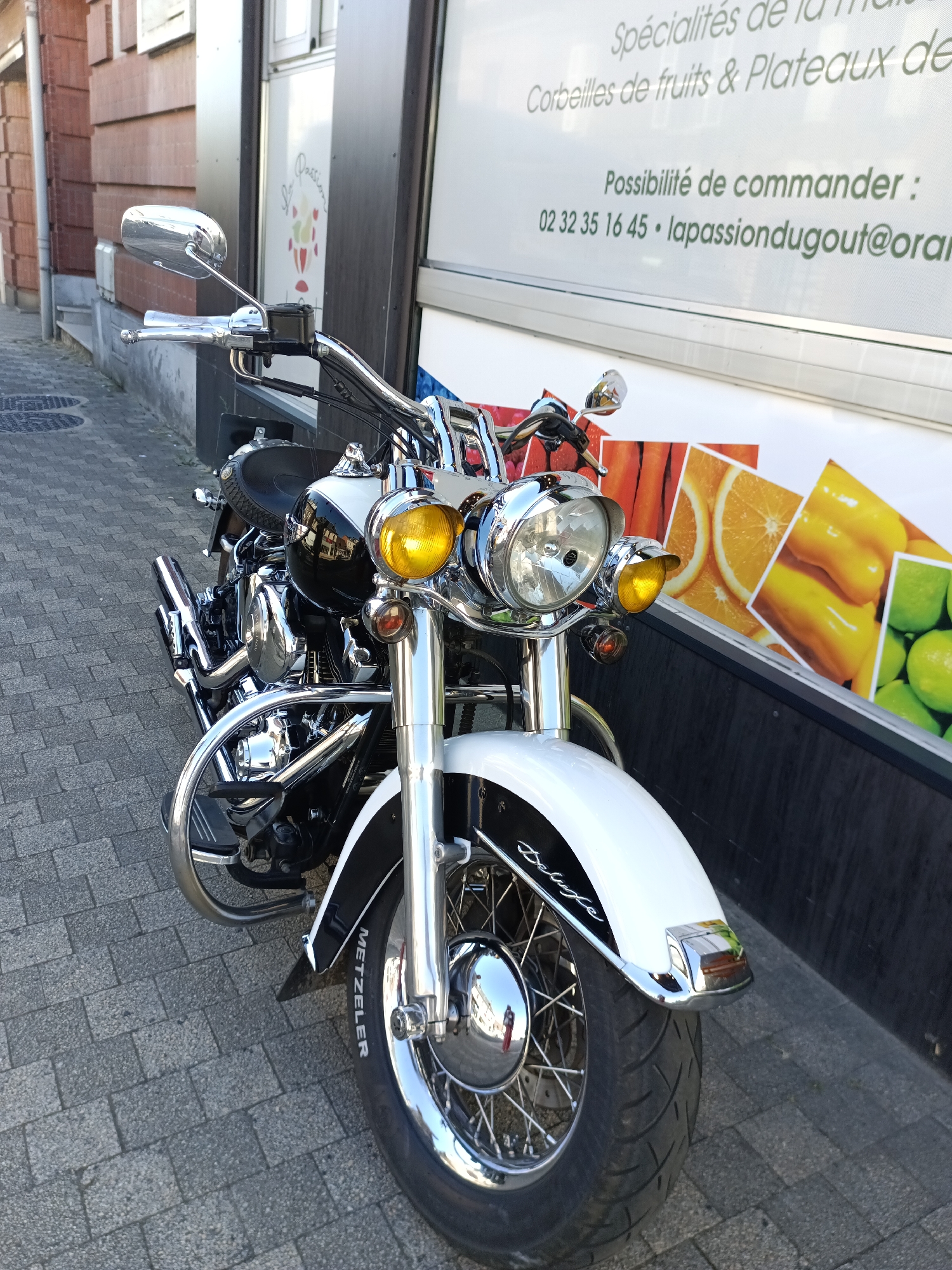 A parked black and white motorcycle is positioned on a brick sidewalk in front of a building in Le Neubourg, France. The motorcycle features extensive chrome detailing, including the handlebars, front forks, engine components, and exhaust pipes. Prominent yellow headlights and turn signals are visible. The front fender is white with black accents and the word "Deluxe" written in script. The brand name "METZELER" is visible on the front tire. Behind the motorcycle, a building facade displays large graphics depicting fruits and vegetables such as oranges, peppers, and limes. Text on the building includes "Spécialités de la mais...", "Corbeilles de fruits & Plateaux de...", and "Possibilité de commander : 02 32 35 16 45 • lapassiondugout@orangen..." The building appears to be a commercial establishment. The sidewalk is paved with interlocking bricks. The overall scene depicts an urban street environment.