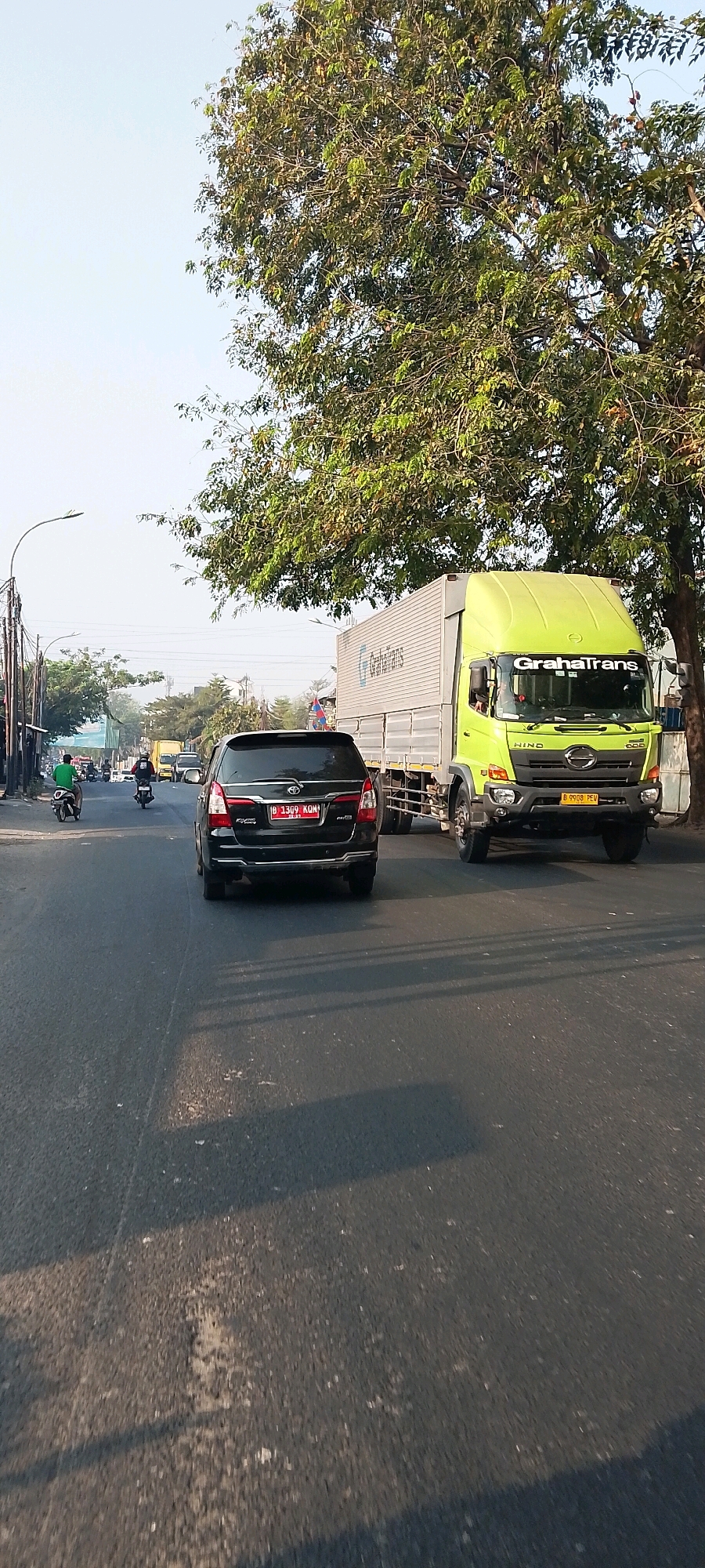 In a medium-shot, an urban scene in Bekasi, Indonesia, unfolds under a bright sky, with a large, leafy tree dominating the upper right quadrant. A paved road, partially shaded by the tree's canopy casting long shadows, stretches towards the horizon. On the road, a black Toyota Innova SUV is in the foreground, its license plate reading "B 1399 KQM". To the right of the SUV is a bright green Hino truck with a white cargo container, emblazoned with "Graha Trans". The truck's license plate reads "B 9908 PEW". Further down the road, several motorcycles are visible. One motorcycle with a rider wearing a green shirt is in the left lane, while another motorcycle with a rider in a dark jacket is further back. In the distance, other vehicles and buildings line the roadside, with streetlights and power lines adding to the urban landscape. The overall impression is one of a typical daytime traffic scene in a bustling Indonesian city.