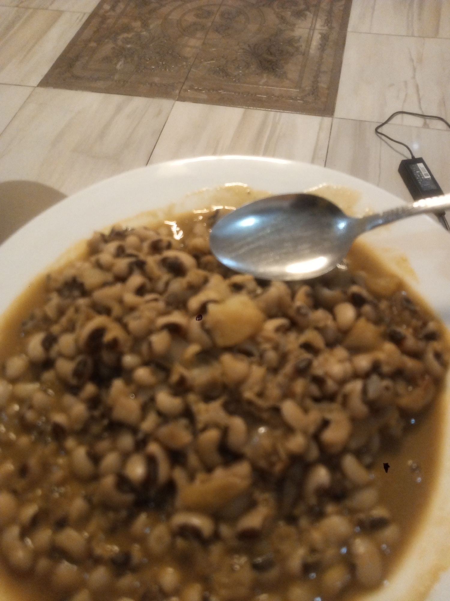 A medium-close shot depicts a plate of stewed black-eyed peas with a spoon. The plate is white. The stew appears to have a light brown gravy. A silver-colored spoon rests on the stew. In the background, out of focus, is a floor composed of light-colored tiles, some of which have a brown patterned design. A black electronic device with a cord is partially visible. This is likely located in a residence or eating establishment in Gwarinpa, Nigeria. There are no people visible.