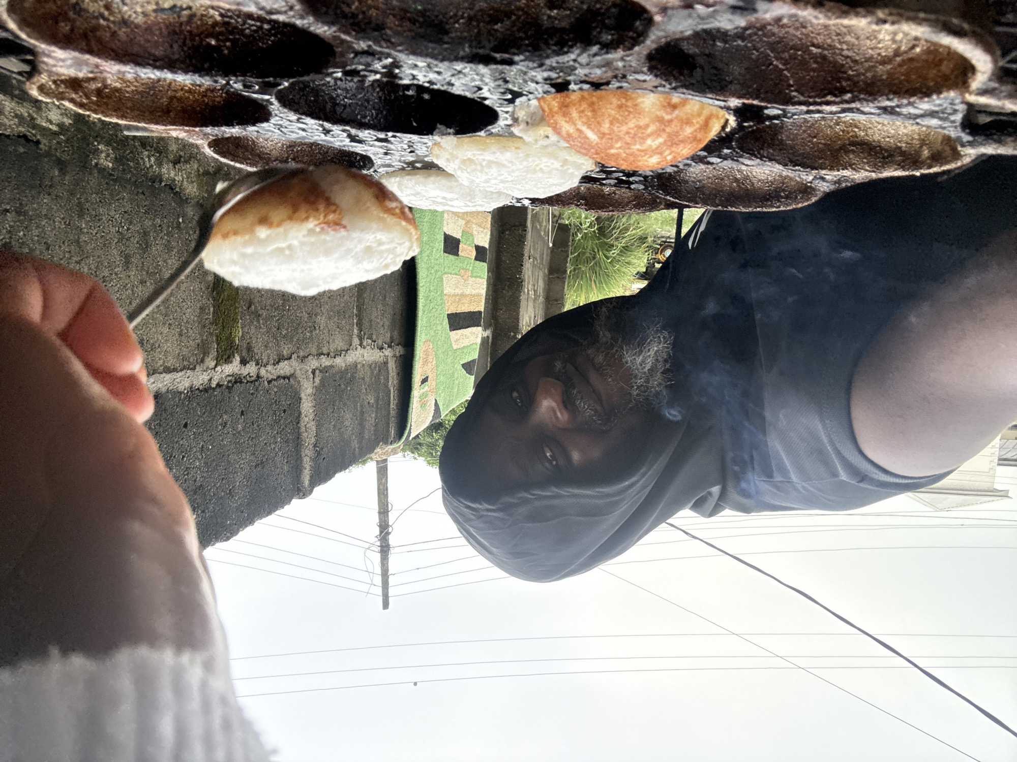 A man wearing a dark grey hooded shirt and a grey tank top is cooking Akara in Ado, Nigeria. He uses a metal utensil to remove a puff of Akara from a dark-colored, multi-cupped griddle. The griddle sits atop a concrete block wall. A green, black, and beige patterned rug hangs vertically next to the wall. Power lines stretch across a bright, overcast sky.