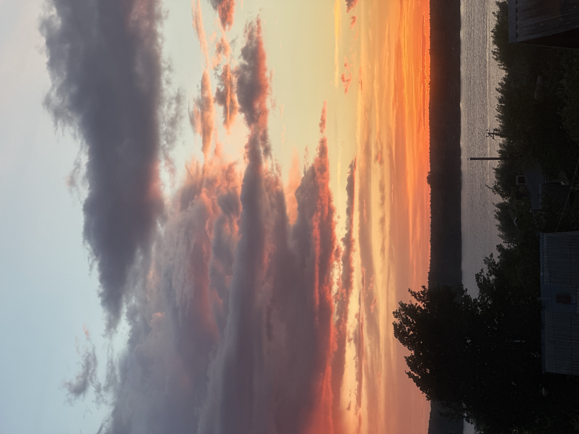The image shows a sunset over a body of water, possibly the Kama River based on the location. The sky is filled with colorful clouds, ranging from dark grays to vibrant oranges and pinks. The colors are particularly intense near the horizon, suggesting the sun is setting. In the foreground, there are silhouettes of trees and buildings, which appear dark against the bright sky. A transmission line with visible pole is also visible. The setting appears to be a residential area near the river.