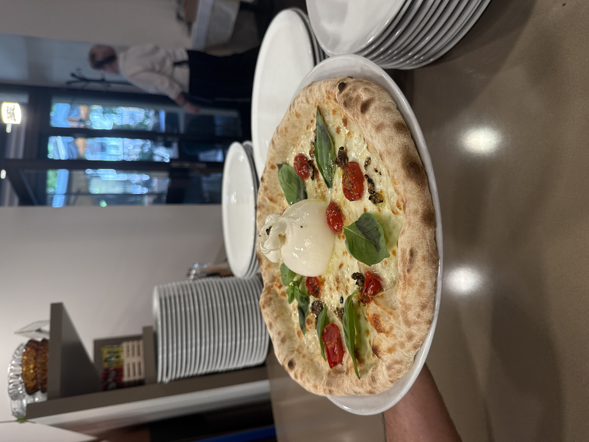 An overhead shot shows a pizza on a white plate held by a person's arm. The pizza has a thick crust and is topped with cheese, cherry tomatoes, basil leaves, and a large dollop of burrata cheese. In the background, stacks of white plates are visible on a shelf and on a table. A woman in a white uniform is standing in the background. The scene appears to be inside a restaurant in Trento, Italy, as suggested by the ambiance and the style of the food.