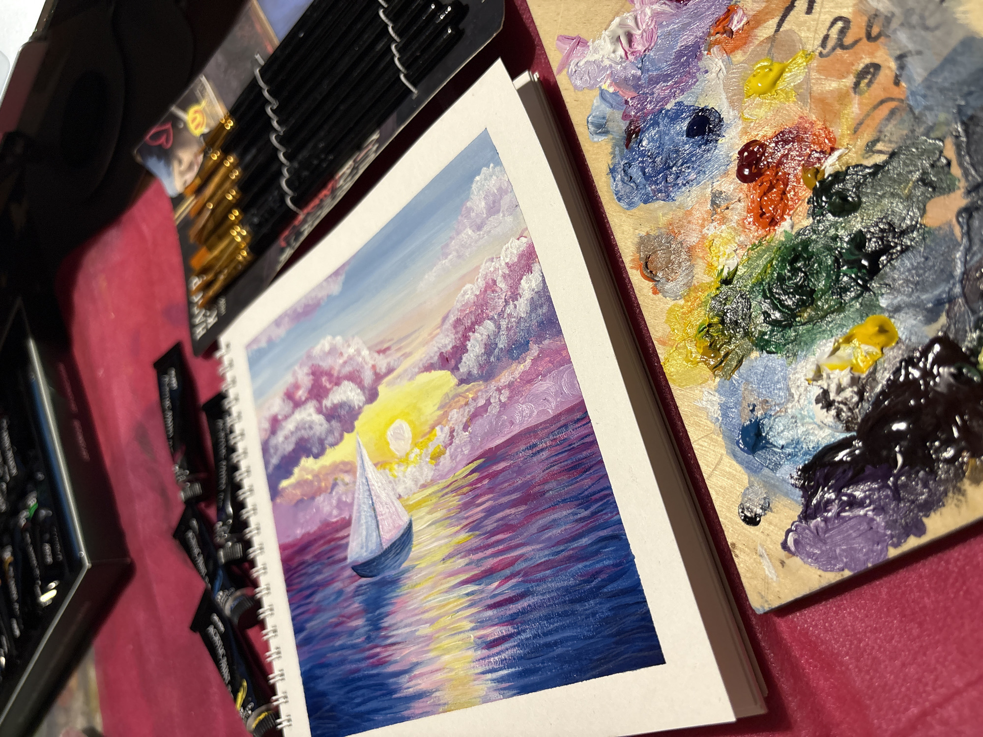 The scene is set in Perm, Russia, depicting an indoor art workspace with various supplies. In the center, a white spiral-bound sketchbook displays a vibrant, vertically oriented acrylic painting. The painting features a stylized boat or craft with a triangular, light blue and pink sail-like structure. The background of the painting depicts an abstract sky with a bright yellow light source and textured pink and white clouds on the right, transitioning to lighter blue at the top left. The lower half of the painting shows water rendered with prominent, jagged vertical brushstrokes in shades of deep blue, purple, pink, and yellow, reflecting the sky.
To the immediate left of the sketchbook, several black tubes of acrylic paint are visible, with "ACRYLIC PAINT" clearly legible on one tube. To the right of the sketchbook, a package of new or unused paintbrushes with black handles and gold ferrules is secured in clear plastic packaging, which also displays a small graphic of a person with a heart and a smiley face.
In the lower right foreground, a wooden artist's palette is covered with numerous distinct blobs of acrylic paint in various colors, including dark purple, black, white, light blue, dark blue, green, orange, red, yellow, and pink, some of which are partially mixed. The entire setup rests on a textured, vibrant magenta or pink fabric surface. The background of the image is dark and out of focus, containing indistinct shapes, possibly other art supply containers.