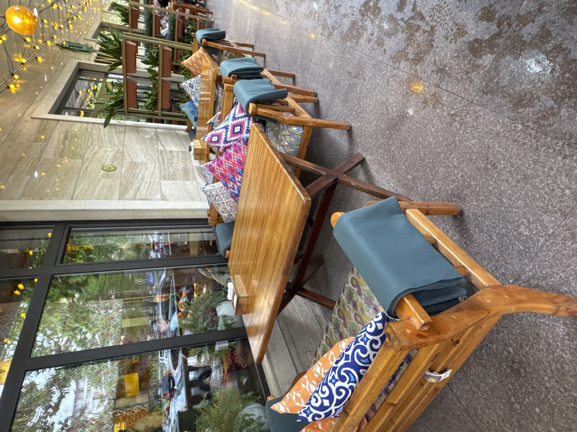 This image presents an outdoor seating area of a cafe or restaurant, likely situated along a sidewalk or promenade in Bishkek City, Kyrgyzstan. The perspective is angled, showcasing a row of wooden tables and chairs.
The furniture includes sturdy wooden tables and chairs, each fitted with teal or dark blue upholstered seat cushions. Many chairs are further accessorized with an assortment of decorative pillows, featuring vibrant patterns such as geometric designs, florals, and abstract motifs in shades of red, pink, orange, blue, and white. The polished wooden surfaces of the furniture reflect ambient light.
To the left, a modern building facade features large, dark-framed windows that reflect the surrounding urban environment, including hints of vehicles, greenery, and distant structures. Above the windows, a light-colored tiled wall is decorated with string lights displaying numerous small golden bulbs, and a prominent, illuminated spherical golden light, contributing to a warm, inviting atmosphere. Below the windows, a lighter, mottled stone or tiled pathway is visible.
To the right, the ground transitions to a darker, speckled grey pavement, which appears wet in several areas, possibly due to recent rain or cleaning, creating subtle reflections. Green foliage from planters is visible both near the building and through the window reflections. The scene is devoid of people, offering a sense of quietude. No discernible text is visible on any objects within the frame.