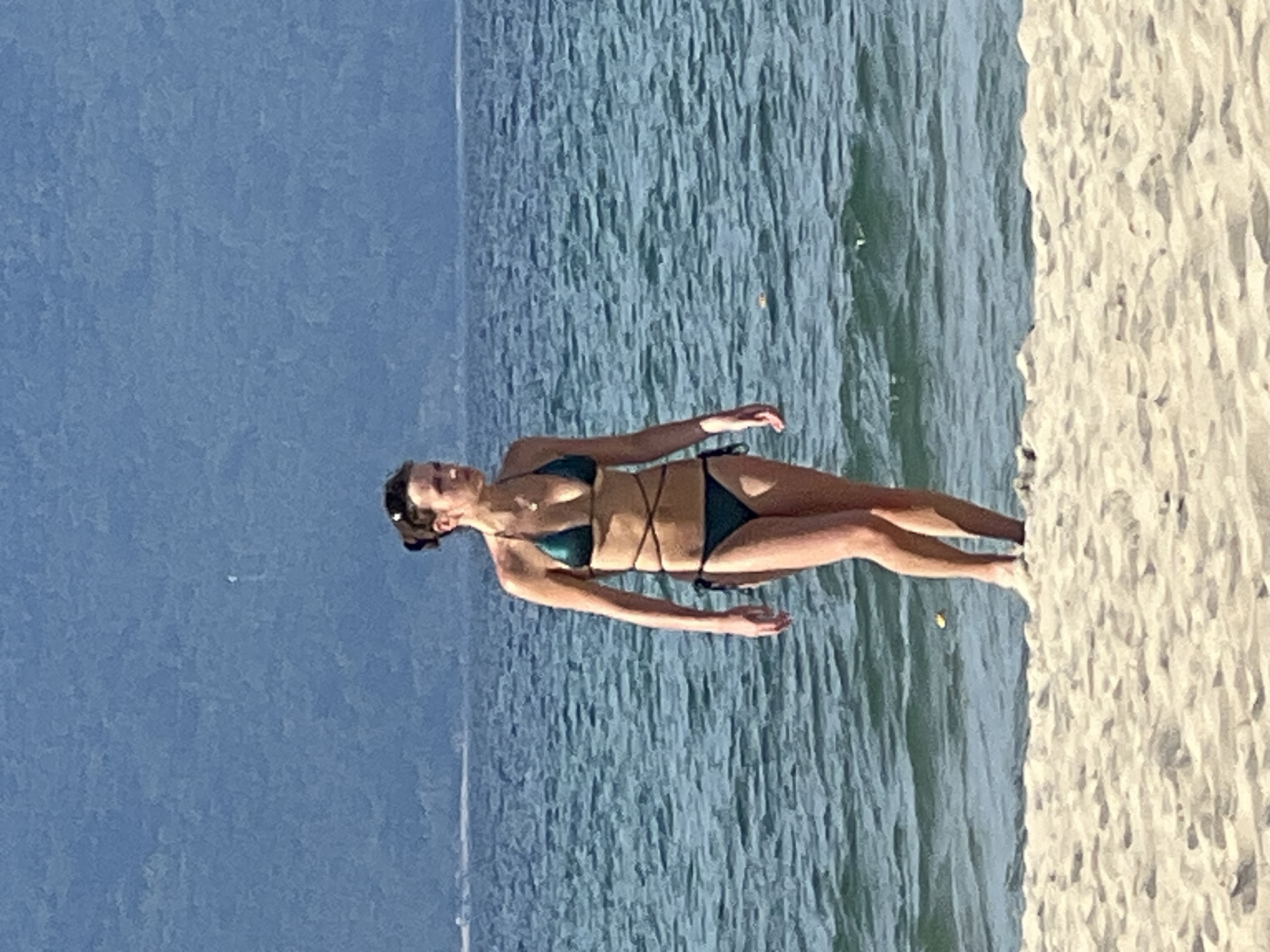 A young woman in a dark green bikini stands at the edge of a sandy beach, with the turquoise ocean stretching out before her and a blurred, mountainous landscape in the background. She has her hair pulled up in a bun and is looking off to her right. The water is slightly choppy, with small waves lapping at the shore. The sand is light in color and appears dry and soft. The scene is characteristic of a beach in the city of Đà Nẵng, Vietnam, a popular tourist destination known for its long sandy beaches and clear blue waters.