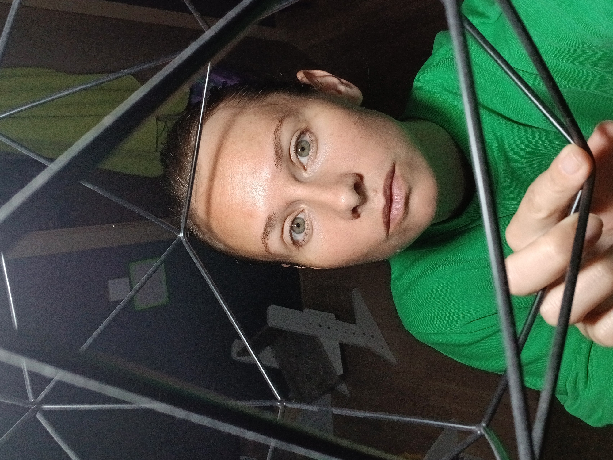 A medium close-up shot captures a woman's face, framed by a geometric metal structure. She has light skin, green eyes, and her hair is pulled back. She is wearing a bright green crewneck sweatshirt. Her left hand is visible, fingers grasping some of the metal bars. The background is dark, with some blurry objects including what appears to be a green towel hanging on a wall, and a white object with holes, possibly a piece of furniture or equipment. The lighting is dim and appears to be from the front, highlighting the woman's face and creating some shadows. The image was taken indoors.
