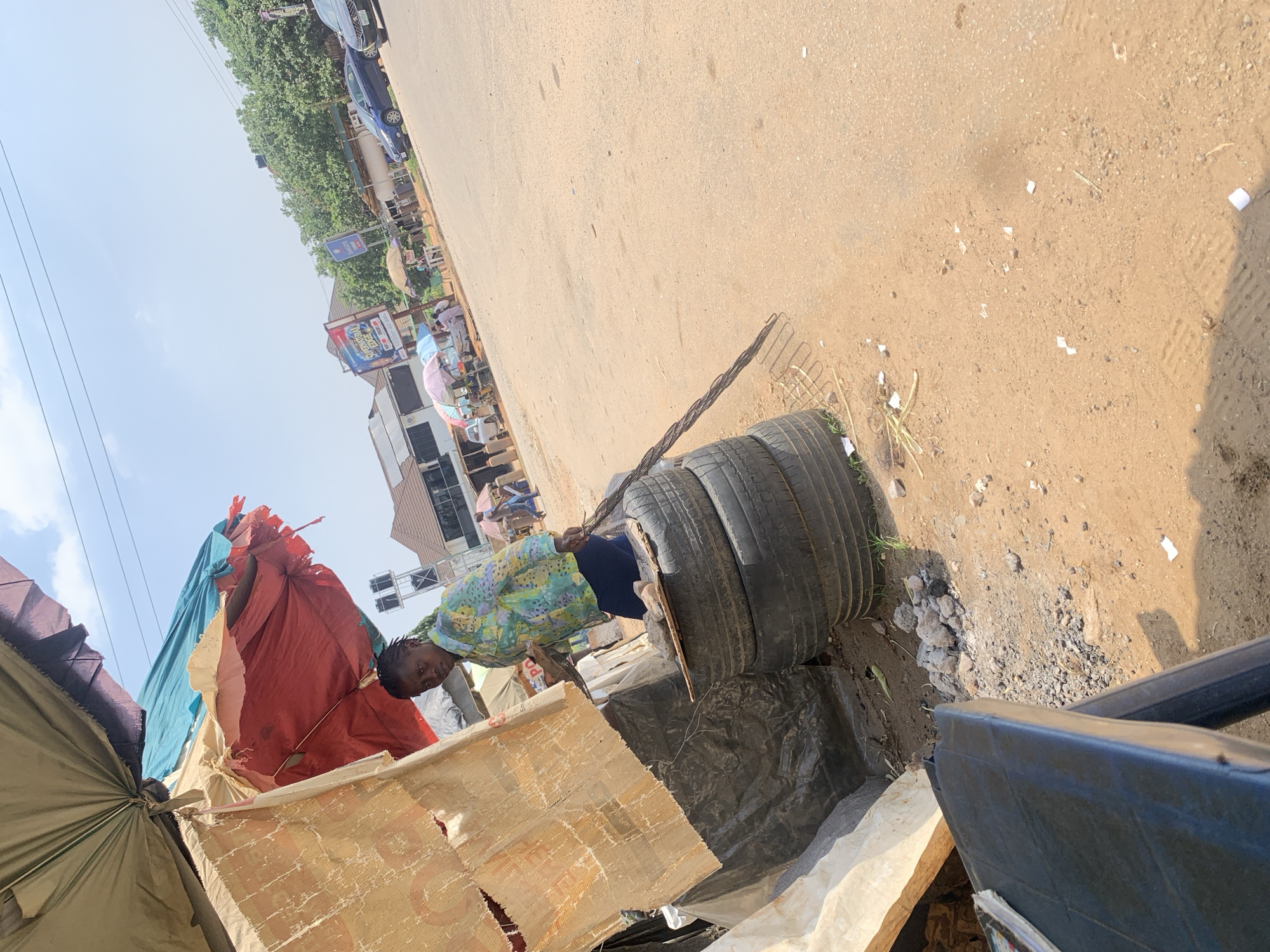 The image shows an outdoor scene in Abuja, Nigeria. A person, possibly a vendor, stands in a makeshift stall with a colorful umbrella providing shade. The stall appears to be constructed from various materials, including what looks like old tires stacked on top of each other, covered with tarpaulin and fabric. On top of the tires, there is a wooden board with some stones. The person is holding a long, slender object, possibly a tool for grilling or cooking. The ground is sandy and dusty, with scattered debris. In the background, there is a street with some cars, buildings, and other market stalls with umbrellas. The sky is bright and clear, indicating a sunny day.