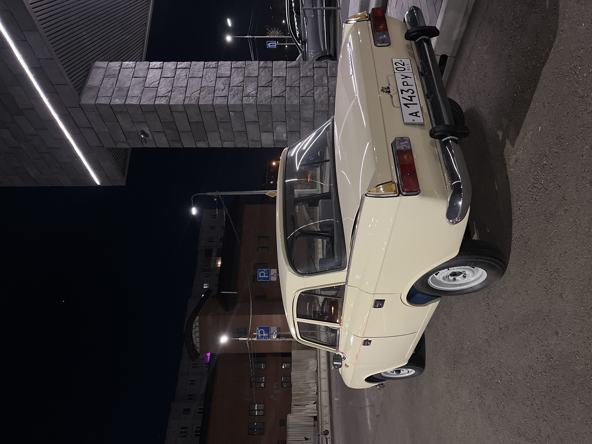 The image depicts a vintage, beige-colored Moskvich car parked in a city street at night. The car is parked near a modern building with an illuminated architectural feature. The license plate on the car reads "A 143 PY 02 RUS". Streetlights and building lights illuminate the scene, and other buildings are visible in the background. Based on the license plate information, the car is registered in Russia. A parking sign is also visible in the background. The specific location appears to be Krasnoyarsk, Russia.