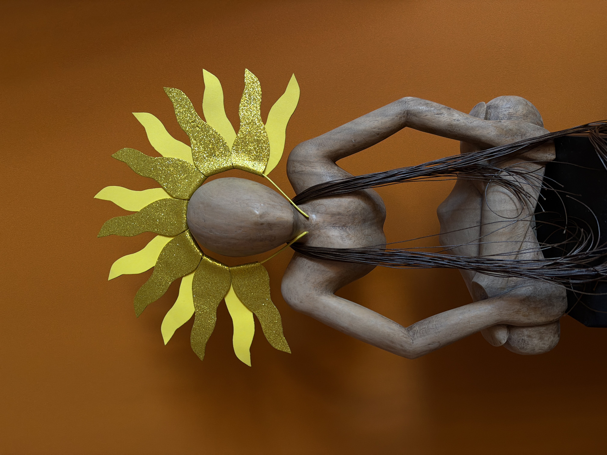 A wooden sculpture of a figure with arms interlocked around itself, wearing a sun-shaped crown, is displayed against a dark orange or brown wall. The sun crown is made of yellow material, with some parts adorned with gold glitter. The figure is attached to a dark object on the right. There is no indication that the image was taken in Khimki, Russia.