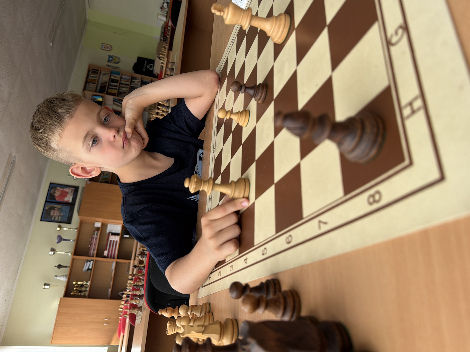 This image shows a young boy, likely of European descent, playing chess. He is seated at a wooden table with a chessboard. The boy has fair skin, light brown hair styled in a buzz cut, and blue eyes. He is wearing a dark blue t-shirt. His left arm is bent, with his hand resting on his chin, suggesting he is deep in thought. His expression is pensive.
The chessboard is set up with chess pieces on it. Both light and dark wooden pieces are visible. The boy's hand is reaching towards a light-colored pawn on the board. The chessboard has alphanumeric notation along its edges. The numbers "6", "7", and "8" are clearly visible.
The background suggests an indoor setting, possibly a classroom or a club room. Shelving units filled with books and trophies are visible behind the boy. Two framed pictures hang on the wall, one depicting two individuals, the other showing what appears to be a sports trophy. The lighting in the room is bright, suggesting it is daytime. No specific time of day or weather conditions are evident. The overall scene conveys a moment of focused concentration during a chess game.