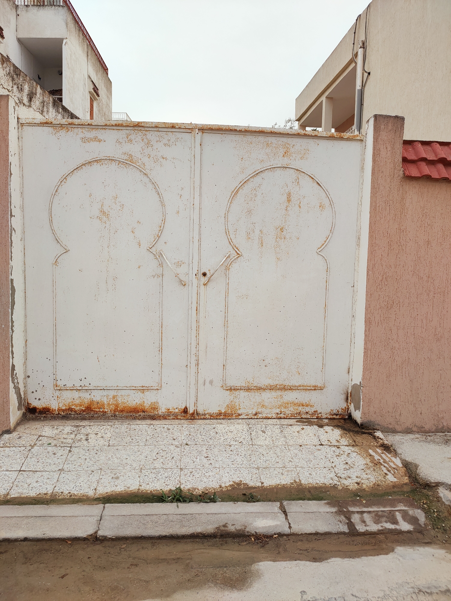 The image depicts a pair of weathered, white metal double gates with arched decorative elements on each panel. Rust streaks are visible along the bottom and sides of the gates, indicating age and exposure to the elements. The gates are set within a rough, pinkish-brown textured wall. To the left and right of the gates, white and beige buildings are partially visible, with a red-tiled roof on the right. The ground in front of the gates is paved with speckled square tiles, and a concrete curb separates it from a muddy street. The overall scene suggests an exterior view of a property entrance in Ariana, Tunisia.