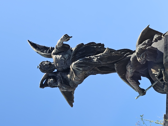 A low-angle shot captures a bronze statue of a winged figure pointing upwards against a clear blue sky. The statue is intricately detailed, with visible folds in the garment and textured wings. To the right of the figure, another figure, possibly cherubic in appearance, grasps what appears to be a staff or rod. The metal has a darkened, aged patina, indicative of outdoor exposure.