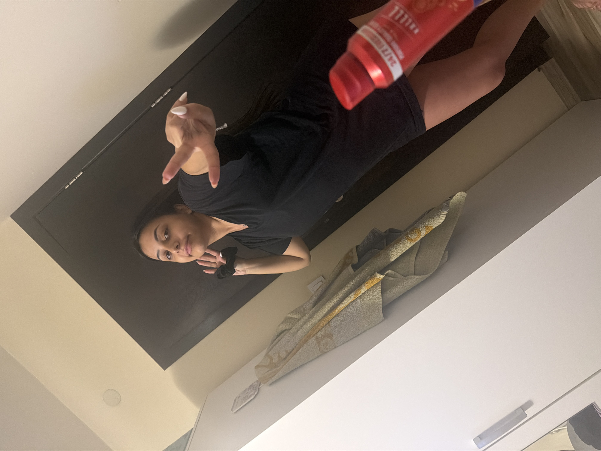 The image captures a young woman, likely in her late teens or early twenties, indoors, possibly in Shumen, Bulgaria. She is positioned towards the camera from a low-angle perspective, and the image itself is rotated. The woman has dark hair and is wearing a black short-sleeved top and dark shorts. Her left hand is raised, showing bent index and pinky fingers, resembling a "rock on" gesture. Her right hand holds a vibrant red plastic bottle, likely containing a personal care product such as shampoo or body wash, with a red cap. Visible text on the bottle includes "24/7" and "DAILY."
The background features a large, dark, flat surface, possibly a wardrobe or cabinet, behind her. To her left, a light-colored wall with a circular fixture (possibly a speaker or light) is visible. Below her, a light-colored, patterned towel, possibly yellow or gold, is draped over a white surface, which appears to be a shelf or another piece of furniture. A small white object, possibly a remote or small container, rests on this white surface. The lighting appears to be artificial indoor illumination. The woman looks towards the camera with a neutral to slightly smiling expression, seemingly posing while holding the product.
