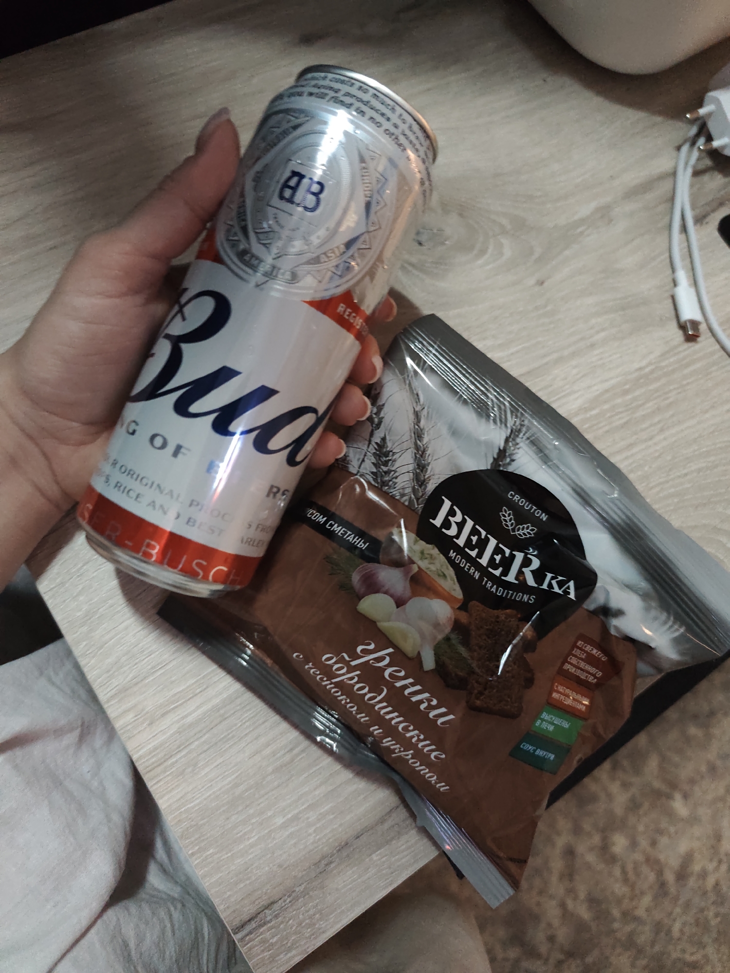 A hand holds a can of Budweiser beer next to a bag of Beerka croutons, borodinsky flavor with garlic and dill. The packaging of the croutons indicates that they are produced in Russia. A portion of a light-colored table is visible. An electrical cord and plug are also visible in the top right corner. The location of the scene is potentially Chaikovsky, Russia, based on the crouton packaging in Russian language.