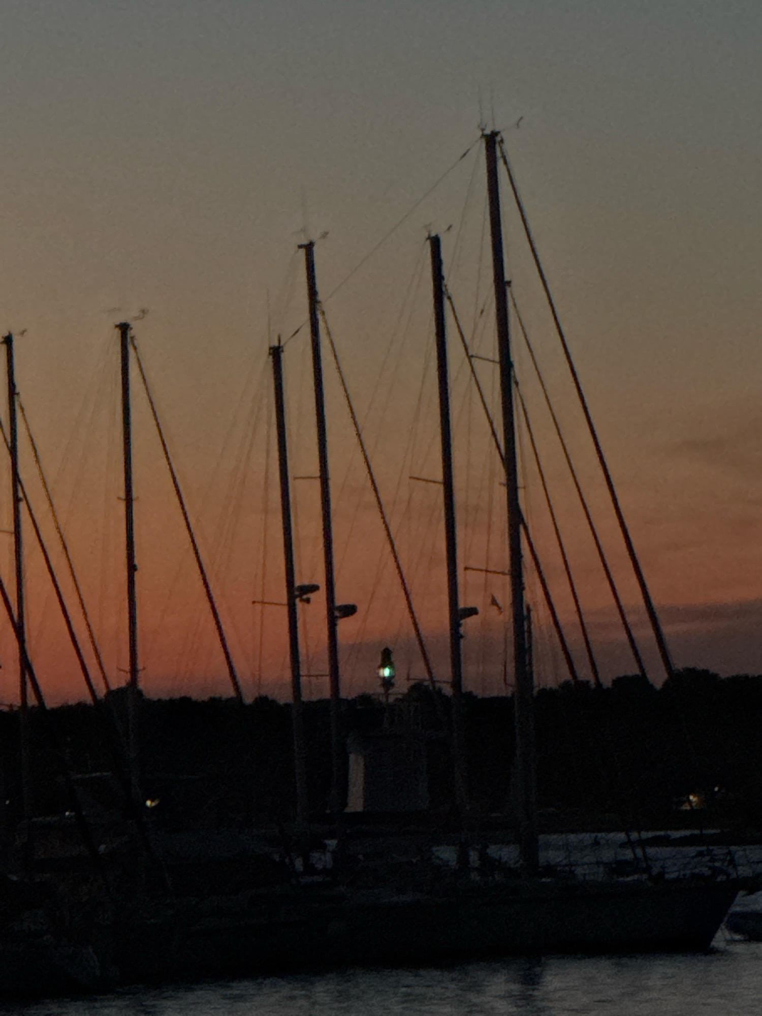 The image depicts a serene twilight scene in the city of Novigrad, Croatia, featuring a marina filled with sailboats. The sky is a gradient of soft oranges and grays, indicative of dusk. Several sailboats, silhouetted against the sky, are docked. Their tall masts and rigging are clearly visible. A lighthouse with a green light is also present, emitting a beacon. The water in the foreground is calm, reflecting the muted colors of the sky and the boats. The overall impression is one of tranquility and the quiet beauty of a coastal evening in Novigrad.