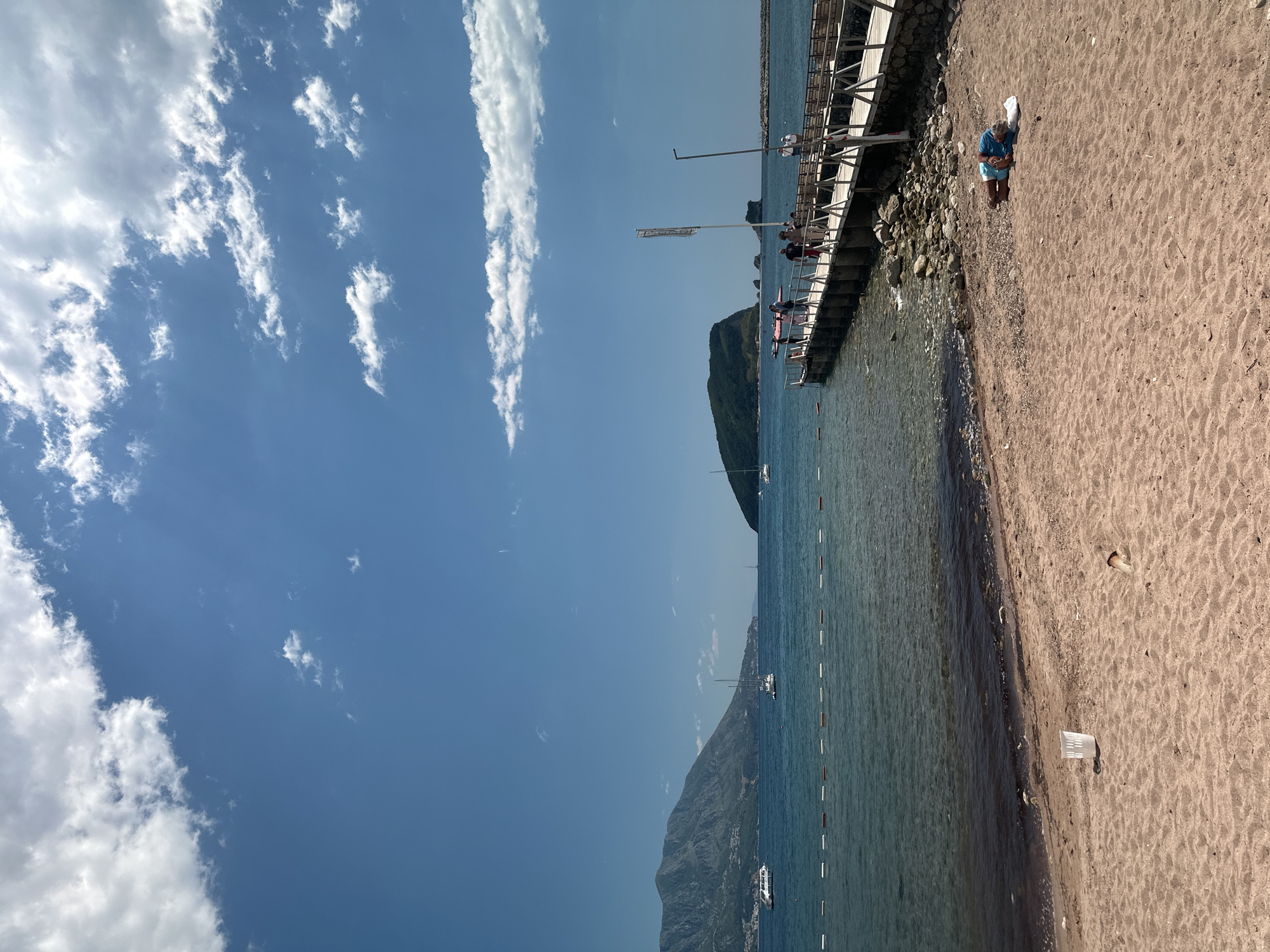 The image shows a beach scene in Budva, Montenegro, with a view of the Adriatic Sea. The beach is sandy, with a small rocky area near a pier. On the pier, there are several people. A woman is sitting on the sand near the pier. In the water, there are several boats. The sky is blue with some white clouds.