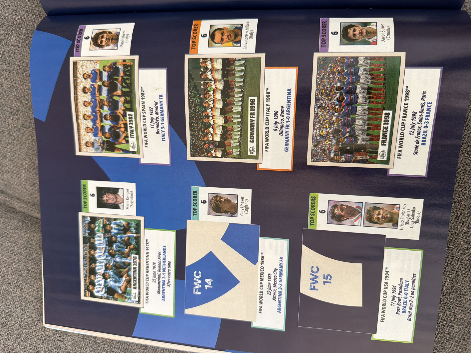 The image is a photograph of an open scrapbook or album page displaying a collection of FIFA World Cup stickers and accompanying information. The album page is dark blue. The stickers are arranged in rows, with each sticker depicting a football team photo or a player's portrait, accompanied by text detailing the event, date, location, and result.
In the top left section, there is a sticker labeled "ARGENTINA 1978" showing the Argentinian team. Next to it, a "TOP SCORER" sticker features a portrait of Mario Kempes (Argentina). Below this, text details the final match of the 1978 World Cup.
To the right of that, a sticker labeled "ITALY 1982" shows the Italian team. Adjacent is a "TOP SCORER" sticker with a portrait of Paolo Rossi (Italy). Below this, text describes the final match of the 1982 World Cup.
In the lower left quadrant, a large sticker with "FWC 14" is partially visible. To its right, a "TOP SCORER" sticker shows a portrait of Gary Lineker (England). Below the "FWC 14" sticker, text details the 1986 World Cup final.
Further down on the right side, a sticker labeled "GERMANY FR 1990" shows the German team. Next to it, a "TOP SCORER" sticker features a portrait of Salvatore Schillaci (Italy). Below the "GERMANY FR 1990" sticker, text describes the 1990 World Cup final.
In the bottom left, another large sticker with "FWC 15" is visible. To its right, a "TOP SCORERS" sticker shows two portraits: Hristo Stoichkov (Bulgaria) and Oleg Salenko (Russia). Below the "FWC 15" sticker, text details the 1994 World Cup final.
In the bottom right, a sticker labeled "FRANCE 1998" shows the French team. Adjacent is a "TOP SCORER" sticker with a portrait of Davor Šuker (Croatia). Below the "FRANCE 1998" sticker, text describes the 1998 World Cup final.
The image does not provide any information or context related to the city of Zlatitrap, Bulgaria.