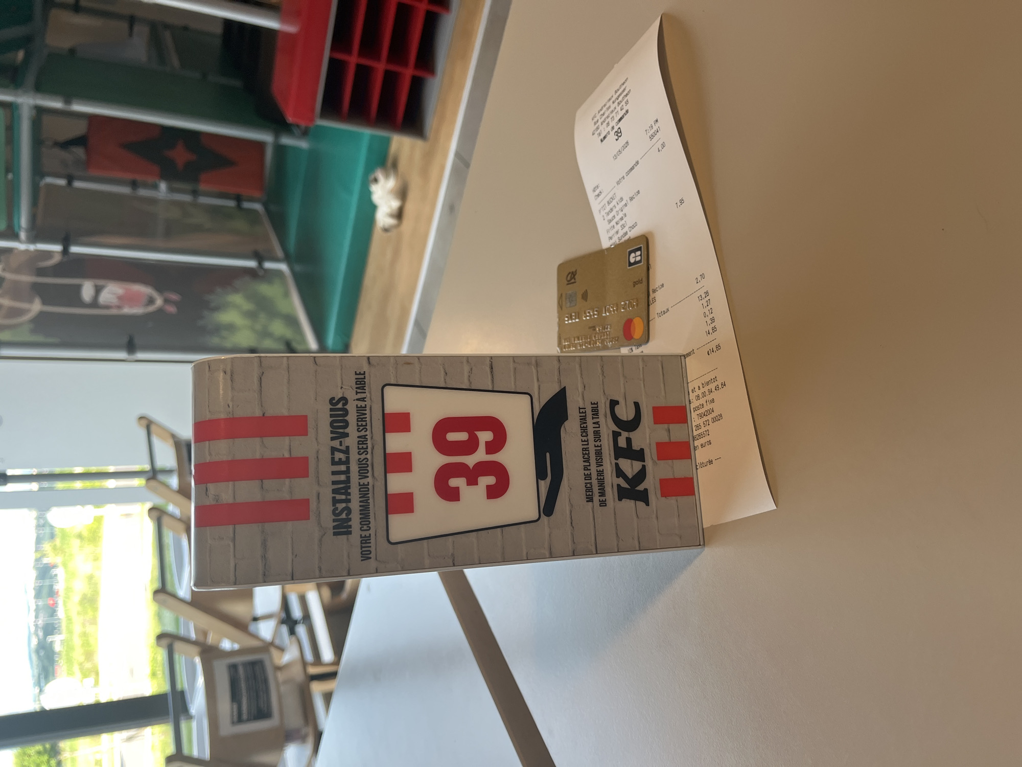 The image shows a table in a KFC restaurant, as indicated by the table sign with the KFC logo and instructions to place the sign visibly for table service. The table number is 39. A receipt and a gold credit card are lying on the table. The receipt shows an order total of €14.65, placed at 7:19 PM on June 13, 2026. The restaurant's phone number is listed as 04 77 31 42 55. The presence of the receipt, credit card, and table sign suggests a customer has recently paid for their meal. Through the window, the location appears to be in Andrézieux-Bouthéon, France. Chairs are seen through the window. Additional tables and booths can be seen in the background of the restaurant, with an object resembling a cat in the background.