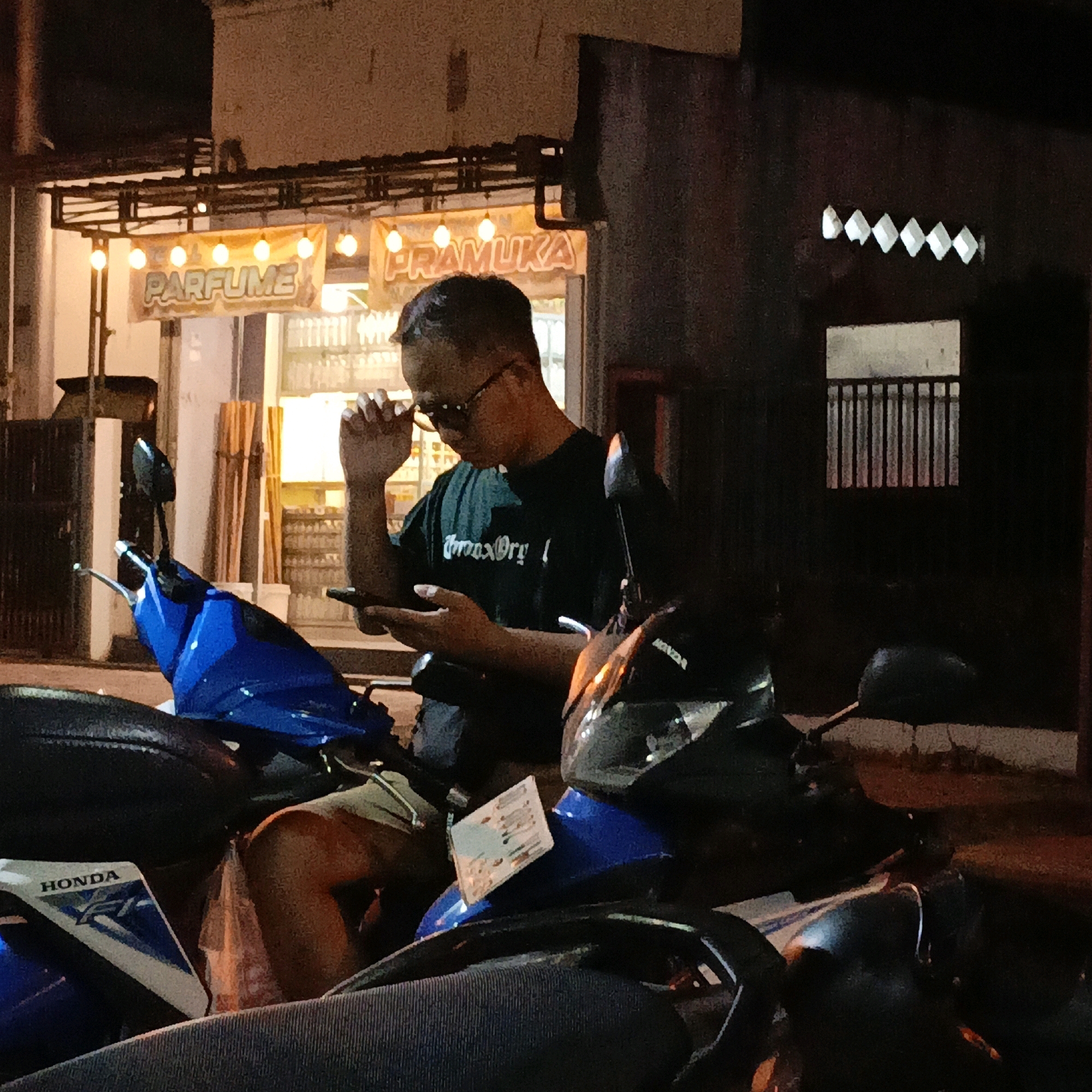 A young man is sitting on a motorcycle at night, in front of a store that sells perfume and other items. He is wearing a dark t-shirt and glasses, and he is looking at his phone. The store is lit with string lights, and there are bottles of products on shelves behind the counter. The man is holding a small bag in his left hand. The motorcycle is blue and has a "HONDA" logo on it. The street is paved and appears to be in a commercial area of Salatiga, Indonesia.