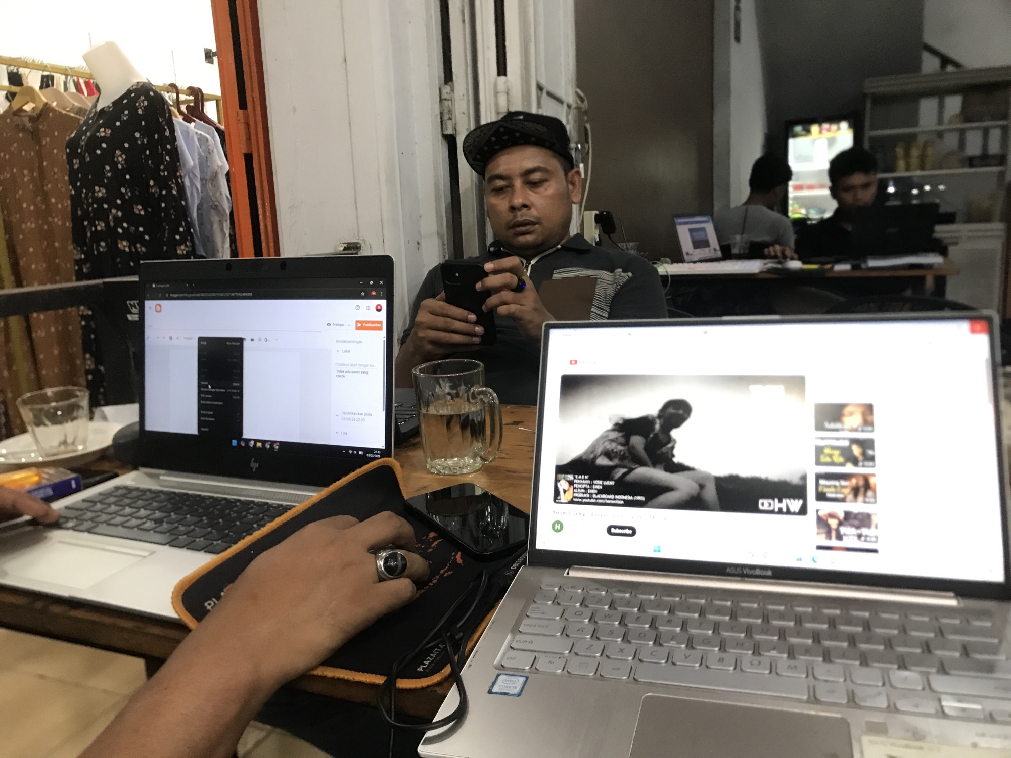 The image depicts an indoor scene with multiple individuals and technological devices. In the foreground, there are two laptops. The laptop on the right displays a YouTube video with the ASUS VivoBook logo visible near the keyboard. To its left, a person's hand rests on a mousepad with a ring visible on the finger. The second laptop, on the left, appears to show a document or blog posting interface. In the center, a man wearing a cap looks at a smartphone. Between the laptops, there's a glass mug and a mobile phone. In the background, two people are visible working at a desk. A refrigerator and a shelf with various items are also visible. Garments are on display to the left of the image, possibly in a commercial setting. The OCR text indicates a detail on the screen of a blog posting site, with the city of Lhokseumawe, Indonesia as the image location.