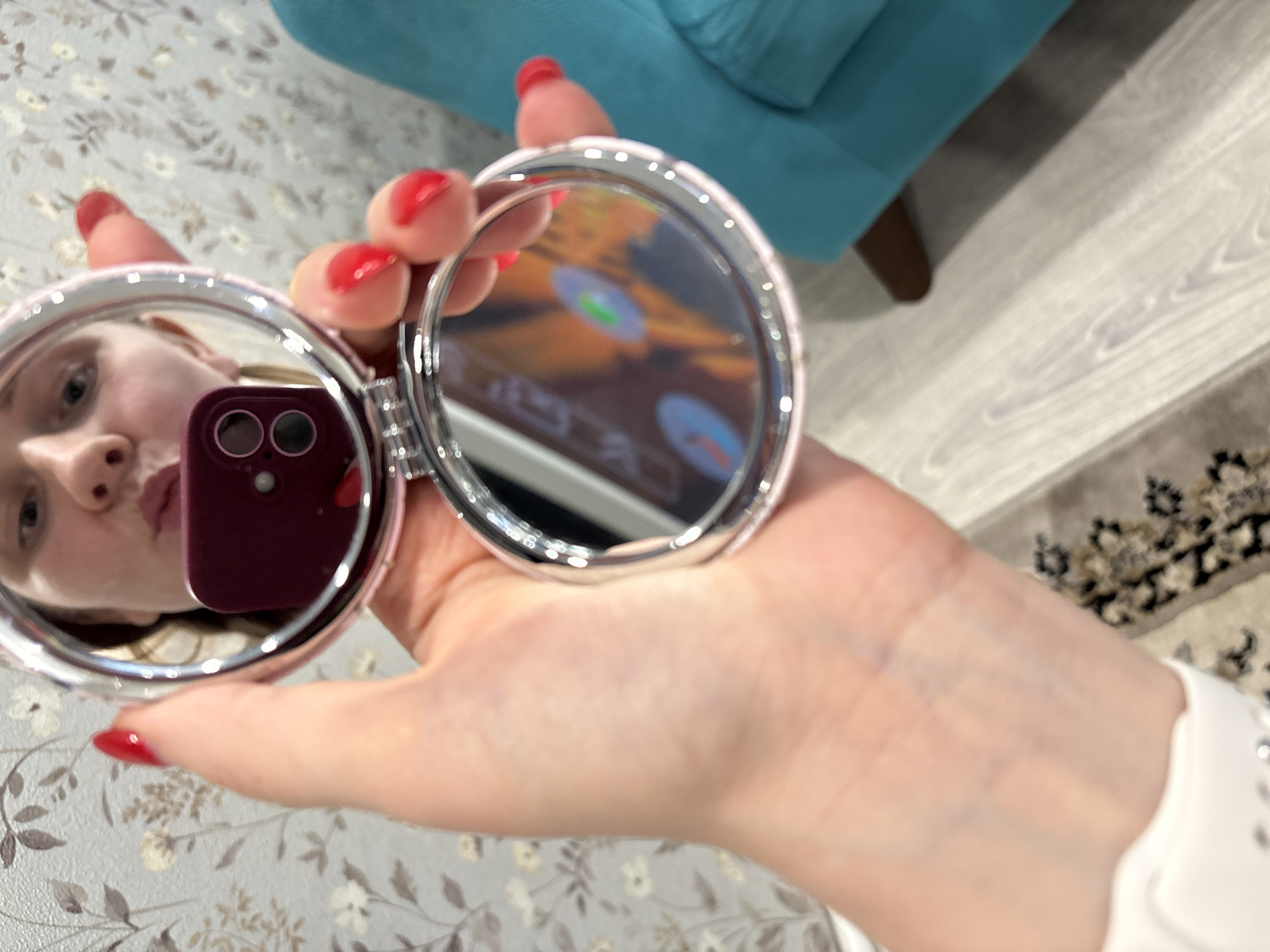 A pale-skinned person with red nail polish is holding a silver-rimmed, dual compact mirror. The person's face and a purple cell phone are reflected in one mirror, while the screen of a larger electronic device is reflected in the other. The background includes floral wallpaper, a blue armchair, and a carpet. The location is Chaikovsky, Russia.