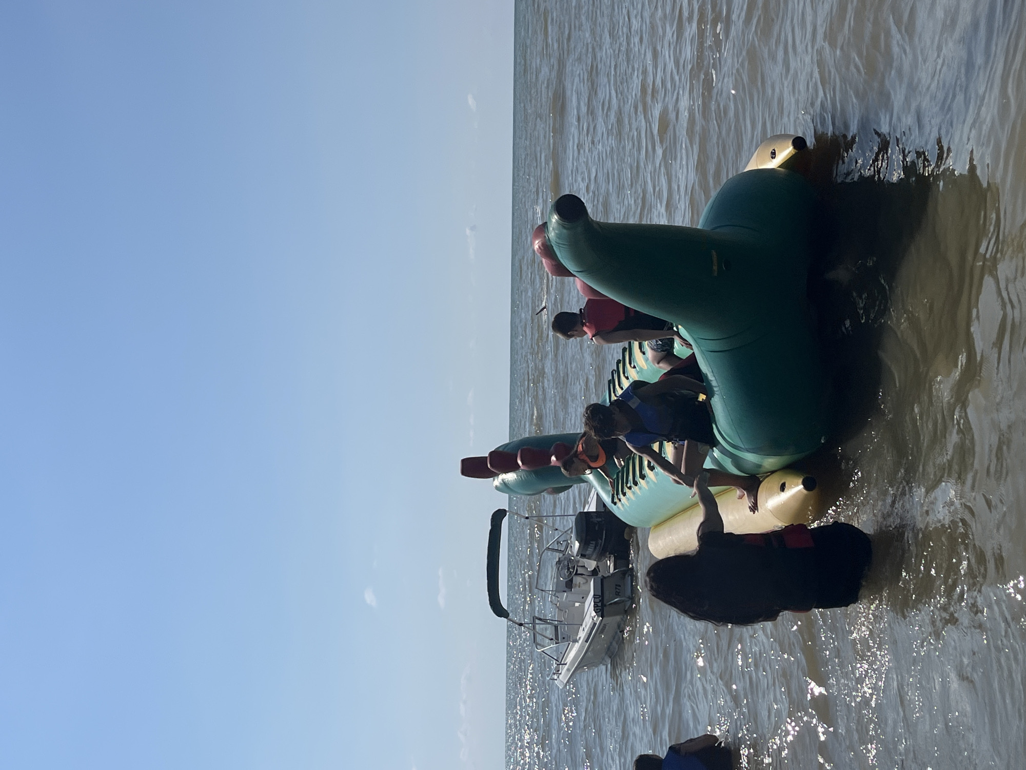 In Ureki, Georgia, on a sunny day, a group of people are gathered on and around an inflatable watercraft shaped like a green dragon. The inflatable is positioned in shallow, dark water, close to the shore, with a white motorboat anchored nearby. Several individuals, including children and adults, are on the inflatable, some wearing life jackets, suggesting an activity related to water sports or recreation. One person is standing in the water, reaching out to the inflatable. The background shows a vast, calm body of water under a clear blue sky, indicating a beach or lake setting.