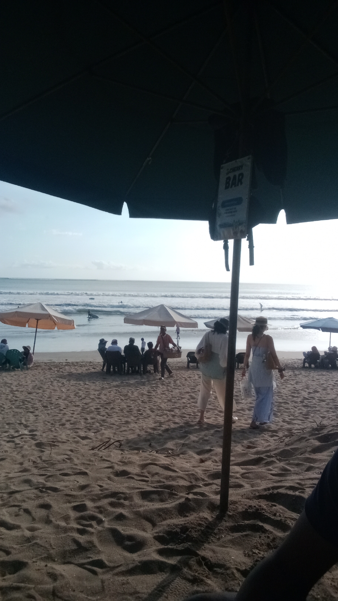 The image depicts a beach scene in Kuta, Indonesia. In the foreground, a dark umbrella partially obscures the view of a sandy beach. A sign on a pole reads "CHERRY BAR". Several beach umbrellas, one orange and two light beige, are set up on the sand closer to the ocean. People are seen seated at tables under some of these umbrellas, and others are walking on the beach. The ocean with gentle waves is visible in the background, and a few individuals appear to be surfing. The sky is bright and clear, suggesting it is daytime. The overall scene portrays a relaxed atmosphere typical of a beach resort area.
