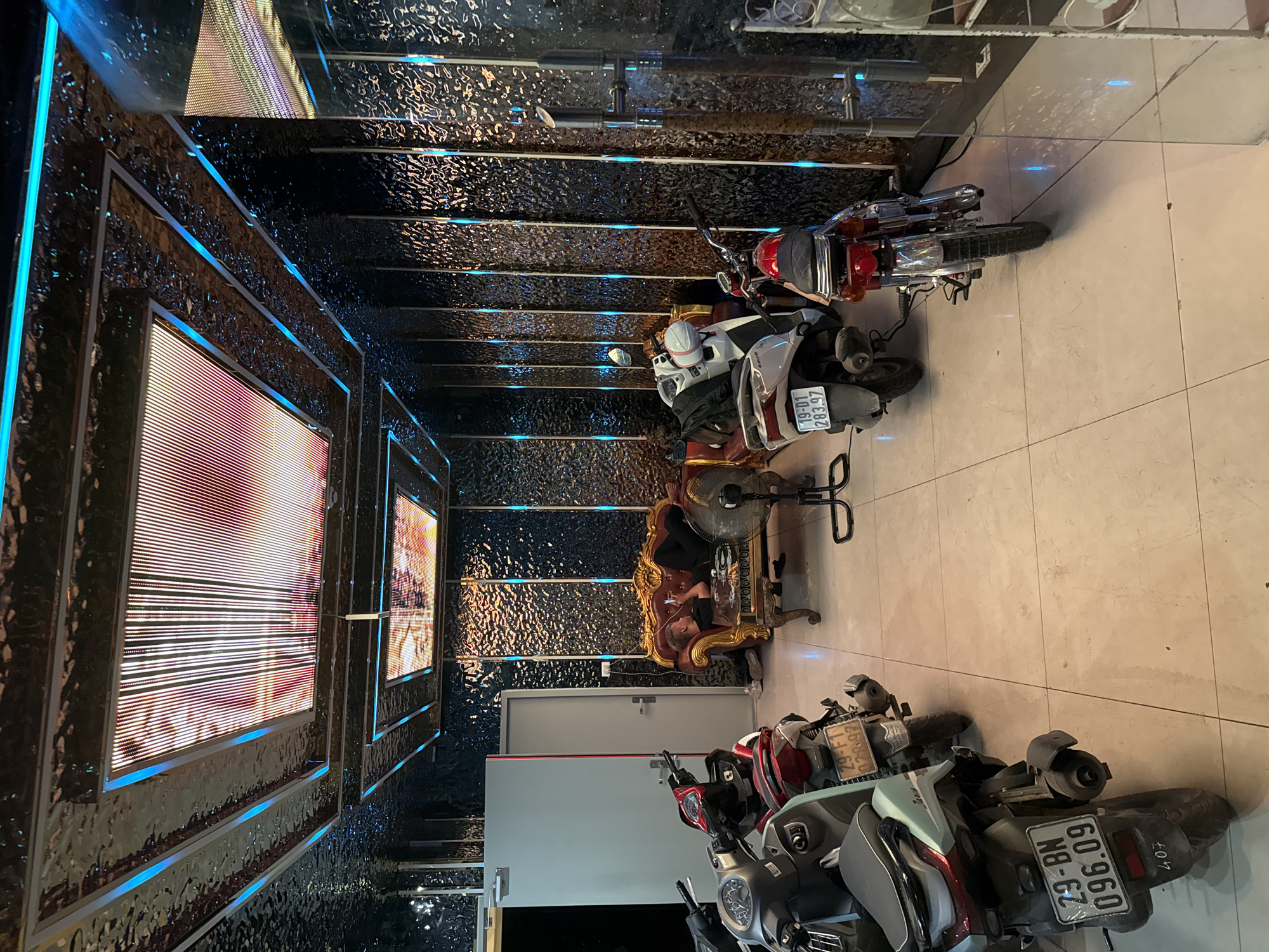 In what appears to be an indoor motorcycle parking area in Hanoi, Vietnam, a person lies on a luxurious golden sofa. The environment is dimly lit with the primary illumination coming from strip lights under the ceiling and along the walls, casting a blue and red glow. The ceiling features recessed panels with light displays. The walls are textured, resembling hammered metal, and are interspersed with vertical blue LED strips. Several motorcycles are parked in the foreground and midground. The person on the sofa is casually dressed and appears to be looking at their phone. A standing fan is positioned near the sofa. The floor is tiled.