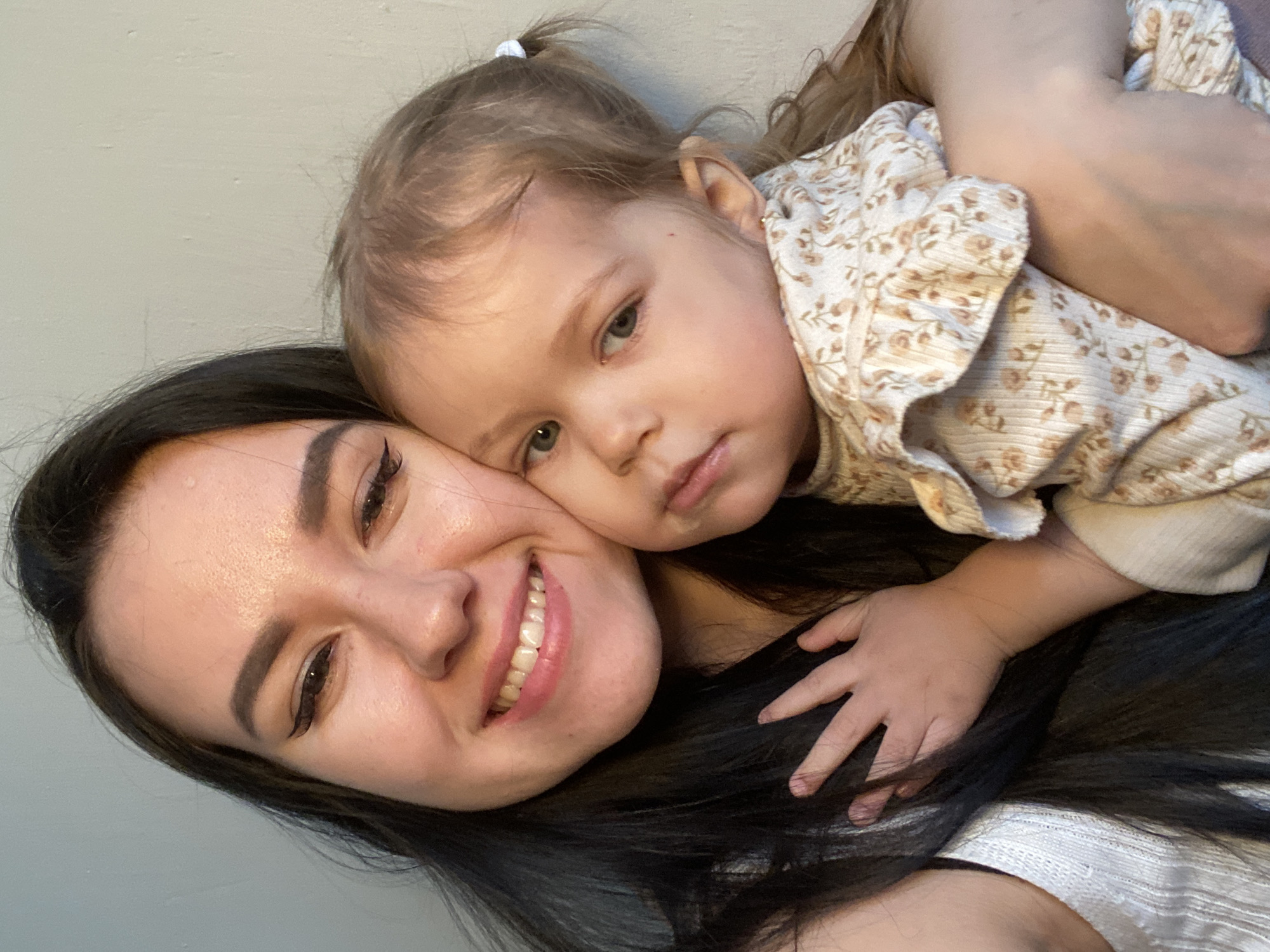 A medium shot depicts a woman and a child, seemingly a mother and daughter, against a neutral gray background. The woman has dark hair, fair skin, and is smiling with visible teeth. She wears eyeliner and has well-defined eyebrows. The child, who is fair-skinned with light brown hair, rests their head on the woman's. The child is wearing a light-colored floral print outfit. The child's hand is resting on the woman's dark hair. The image is rotated, causing a slight tilt. The city and location of the image are not discernible.