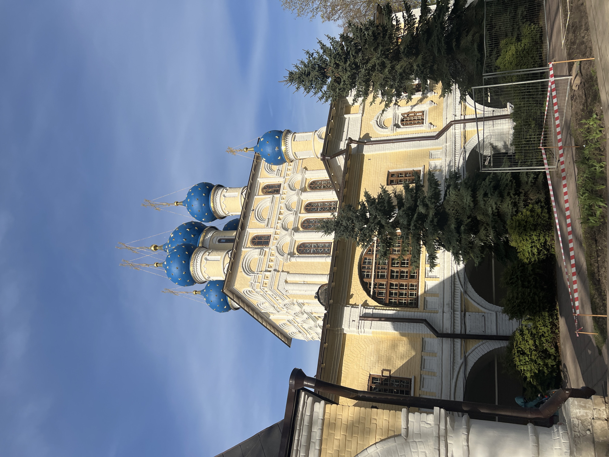 The image shows a partial exterior view of the Church of the Holy Mandylion in Moscow, Russia, photographed during daylight. The church is a primarily yellow building with white architectural details, topped with five blue domes decorated with stars. Evergreen trees partially obscure the facade. A temporary construction fence with red and white caution tape is visible on the right side. The sky is clear and blue.