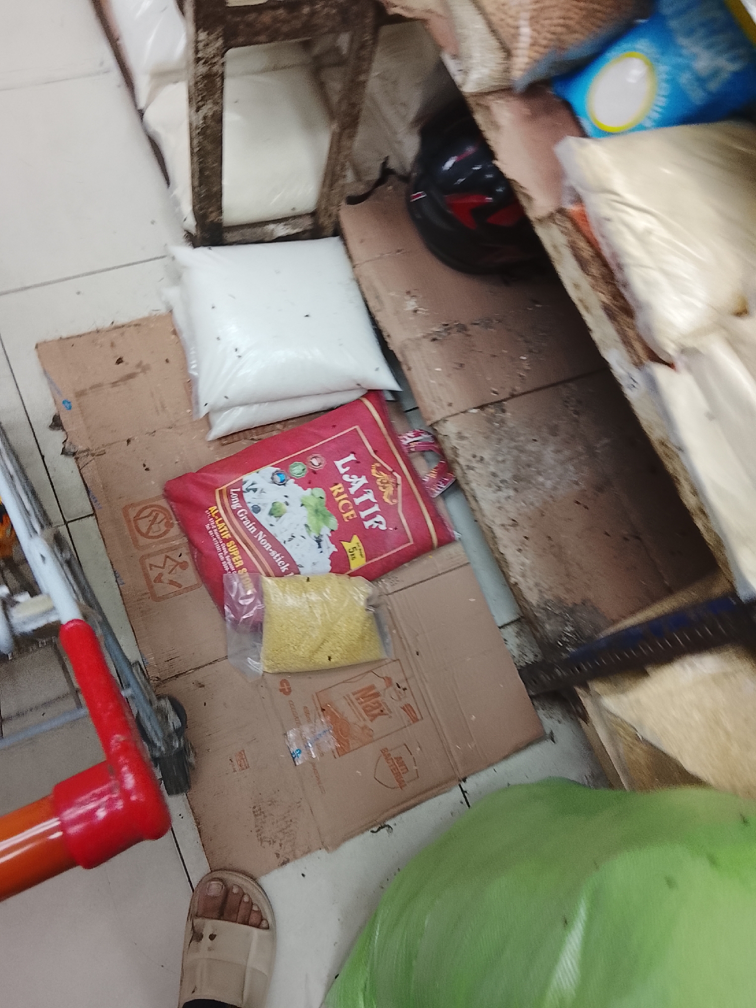 The scene is a low-angle, close-up shot focusing on a segment of a cluttered floor in what appears to be a store or storage area. The setting is potentially in Gulgasht Colony, Pakistan, based on the text on the red "Latif Rice" bag.
The floor is covered with a piece of cardboard serving as a makeshift mat. On the cardboard are several objects: a white plastic bag, a red bag of "Latif Rice", and a small plastic bag filled with yellow grains, likely pulses. A red-handled trolley or dolly is visible on the left edge of the frame. To the lower right, a person's foot clad in a slide sandal is present. A large, green plastic-wrapped object, its contents obscured, occupies a significant portion of the lower-right corner.
The background is blurry. Additional sacks are arranged. A black helmet is slightly visible and more bags and a weathered wooden structure are in the background. The scene suggests a storage or retail environment where food items are stored and moved around. The overall impression is of a crowded and potentially informal setting.