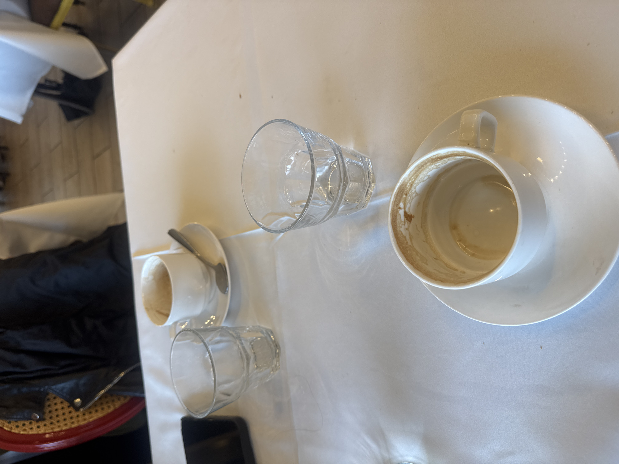 The image captures a tabletop in a café setting in Unknown, Unknown. The table is covered with a white tablecloth, upon which are several used beverage containers. There is an empty white coffee cup with a saucer, showing coffee stains, along with two empty faceted drinking glasses. A second smaller empty coffee cup and saucer is also present, with a spoon resting on the saucer. Adjacent to the table, part of a chair with a red frame and rattan seat is visible, along with a black jacket.