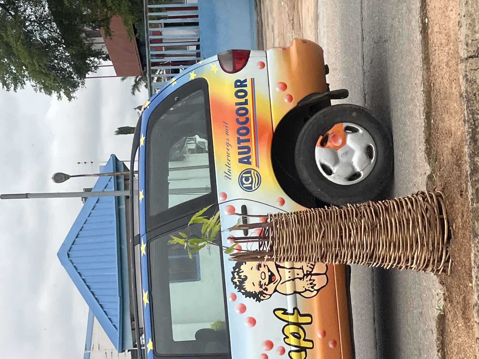 The image shows the side of a colorful van, likely parked on a street in Jalingo, Nigeria. The van is decorated with an orange and yellow gradient and features cartoon drawings of a smiling baby and pink bubbles. The words "Unterwegs mit ICI AUTOCOLOR" are written on the side of the van. A wicker basket with a small plant growing out of it is positioned in front of the van. In the background, a blue building with a triangular roof and a fence can be seen. The sky is overcast.