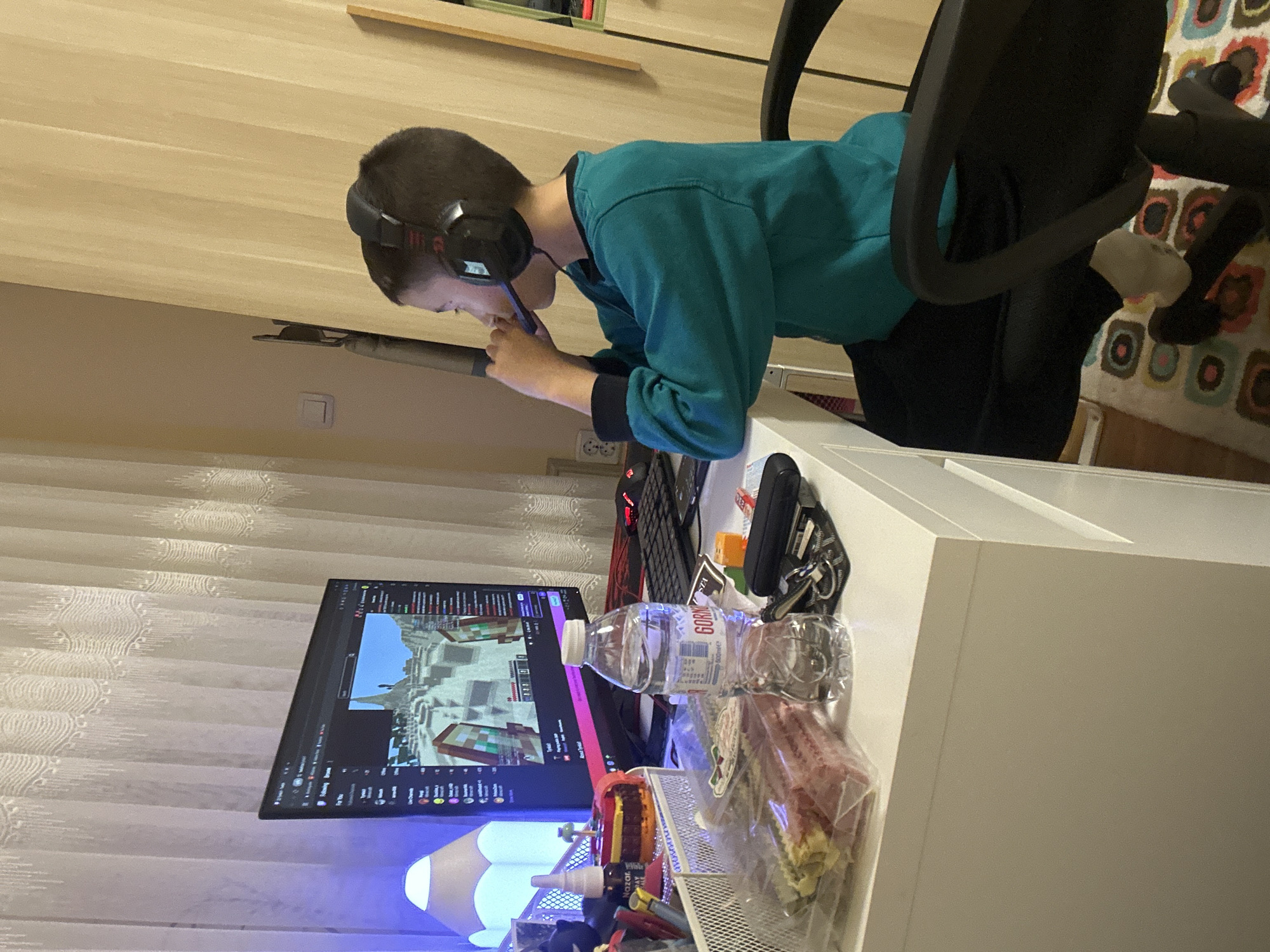 The image displays a young male deeply engaged with his electronic devices at a desk, likely in a home setting within Plovdiv, Bulgaria. He is seated in a black office chair, wearing black headphones and a teal long-sleeved shirt, leaning forward over a white desk as he focuses intently on a smartphone or small tablet held in his hands.
To his left, a computer monitor is active, displaying what appears to be a video game with blocky graphics, strongly resembling Minecraft. The monitor also shows interface elements like "Following," "Browse," and "For You," suggesting he might be watching a stream or video content. A black keyboard with some red illumination and a black mouse are positioned near the monitor.
The desk holds several items, including a clear plastic water bottle with "Gorna Banya" and "600ml" printed on its label, a set of keys on a keyring, and a packaged snack. A small lamp emitting a blue glow and a mesh organizer containing various small items like pens are also visible.
The background features light-colored wooden paneling or furniture and translucent white curtains. A wall outlet, typical of European standards with two round holes and a grounding pin, is present on the wall. A colorful patterned rug with abstract shapes in shades of brown, orange, white, black, and teal is visible beneath the chair. The presence of the "Gorna Banya" water bottle provides a specific location cue consistent with Bulgaria. The scene captures a moment of focused screen time, likely during the evening or an overcast day given the indoor lighting.