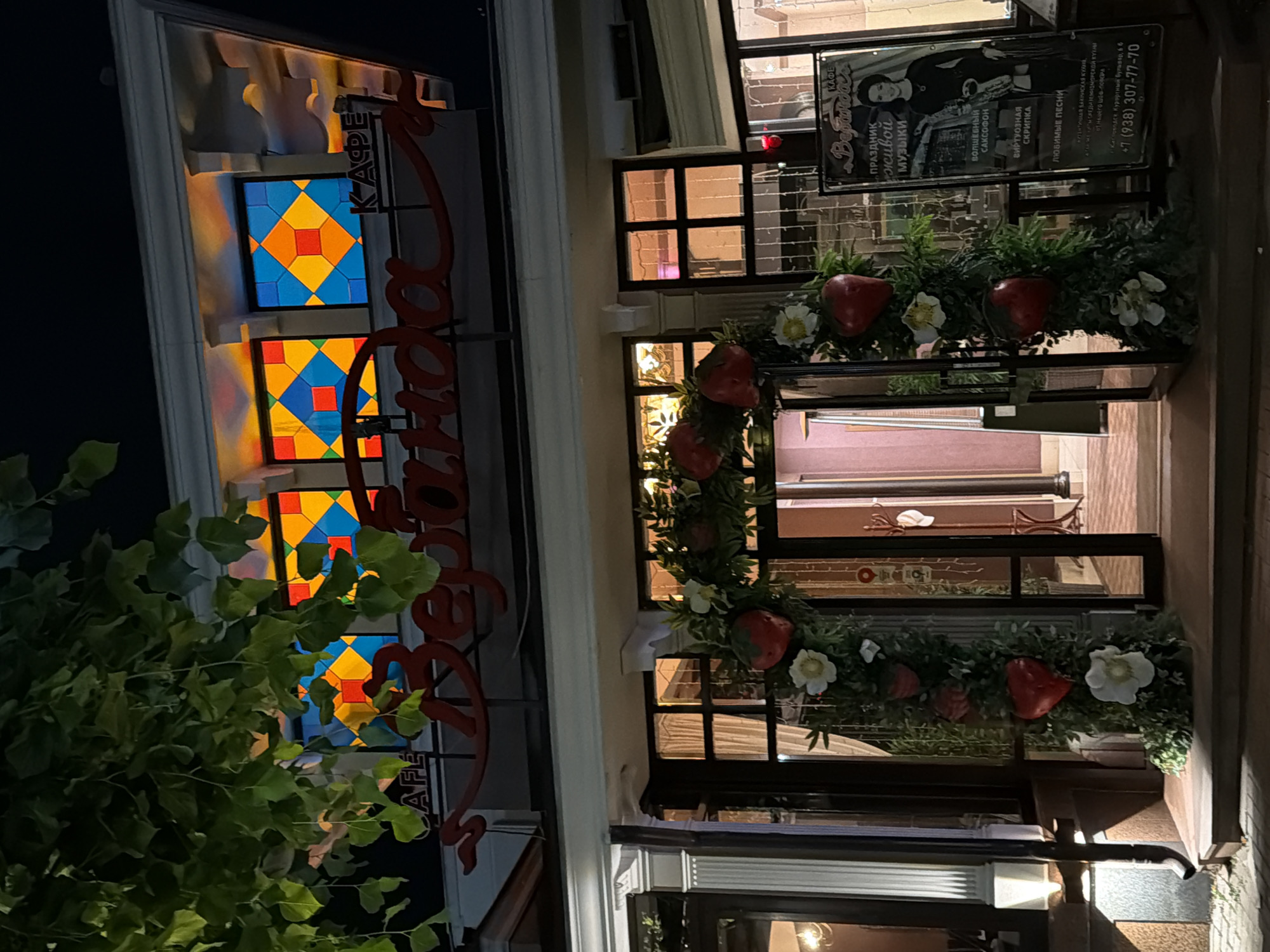 The image shows the entrance to a cafe named "Veranda" in Kislovodsk, Russia. The cafe has a distinctive red sign with white lettering and is decorated with a floral arch featuring large red hearts and white flowers. Above the entrance, there are stained-glass windows with geometric patterns in shades of blue, yellow, and red. To the right of the entrance, a poster displays a woman in black attire and text in Russian, advertising live music, a saxophone, and popular songs. The scene is lit by artificial lights, indicating it is nighttime. The overall impression is of a welcoming establishment, possibly decorated for a festive occasion.