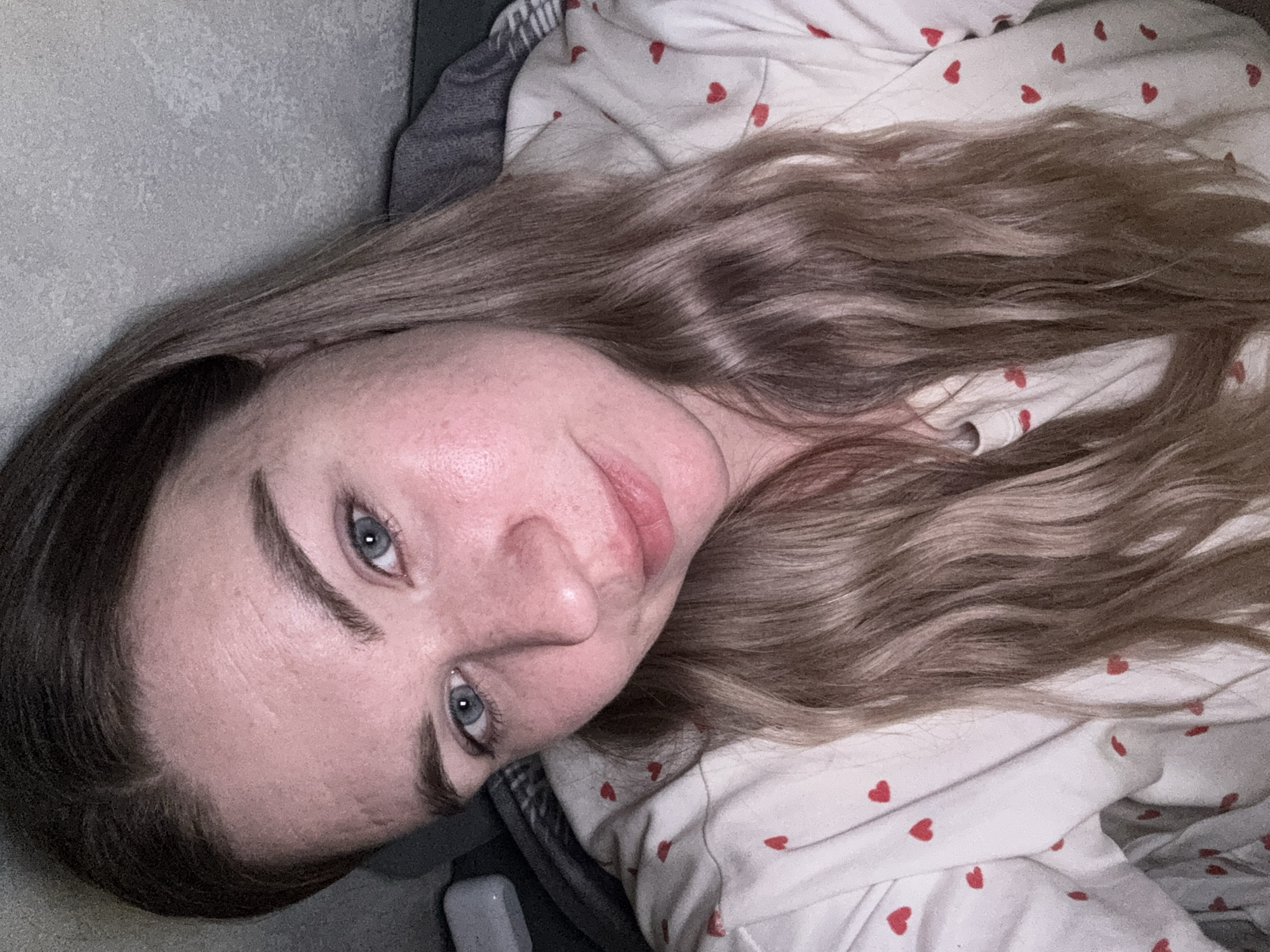A woman with light skin, blue eyes, and light brown wavy hair is lying down, facing the camera. She is wearing a white top with small red hearts. The background includes a grey surface and possibly bedding. The location is not discernible from the image. There is no indication of the city of Chaikovsky, Russia, in the image.
