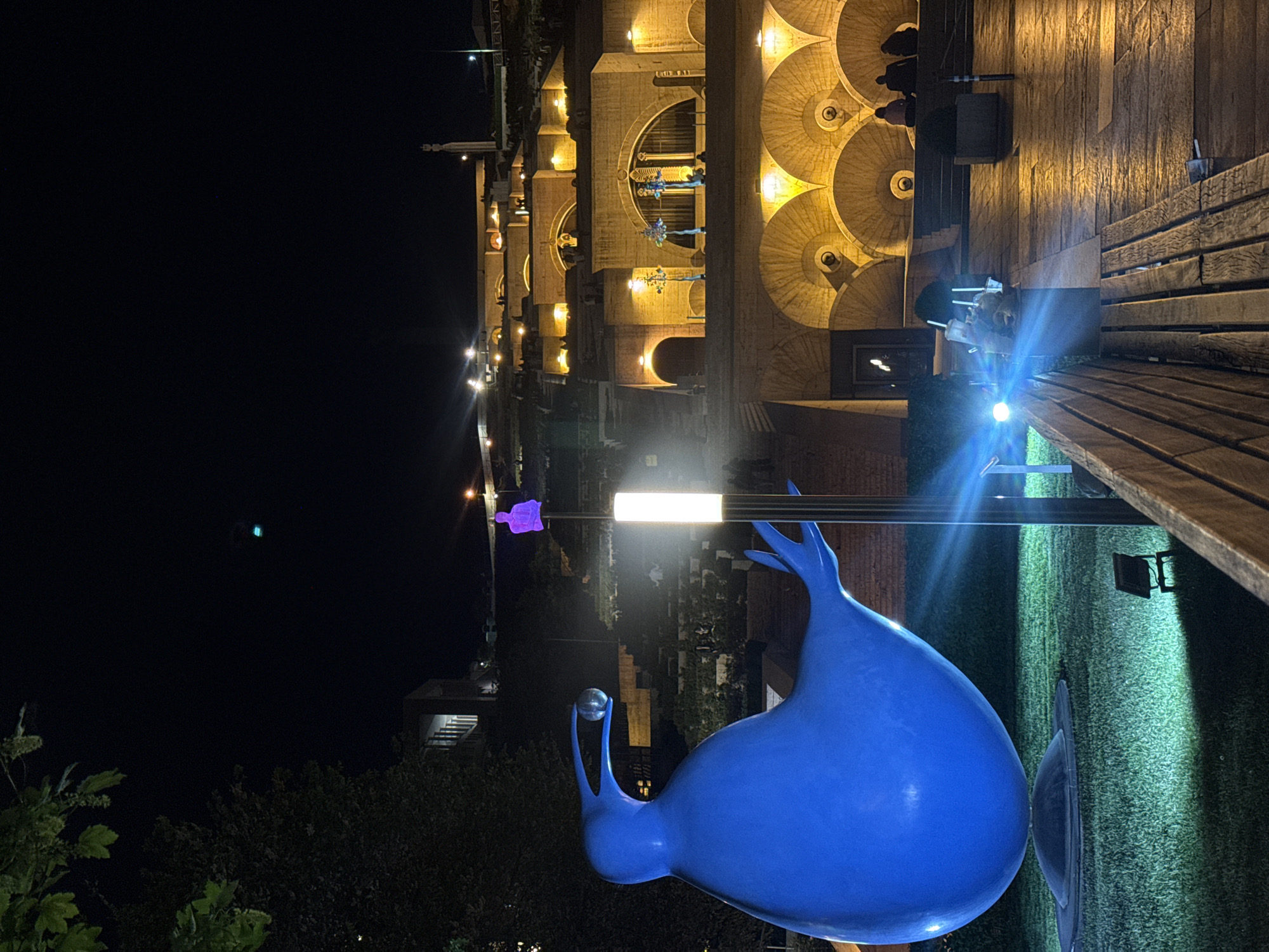The image shows a night scene at the Cascade complex in Yerevan, Armenia. A large, blue sculpture of a whimsical figure with a ball is in the foreground. Behind the sculpture, the illuminated terraces of the Cascade rise up, with rounded archways and decorative elements. A wooden bench is visible to the side. A streetlight illuminates the scene, and other buildings and greenery can be seen in the background. The sky is dark.
