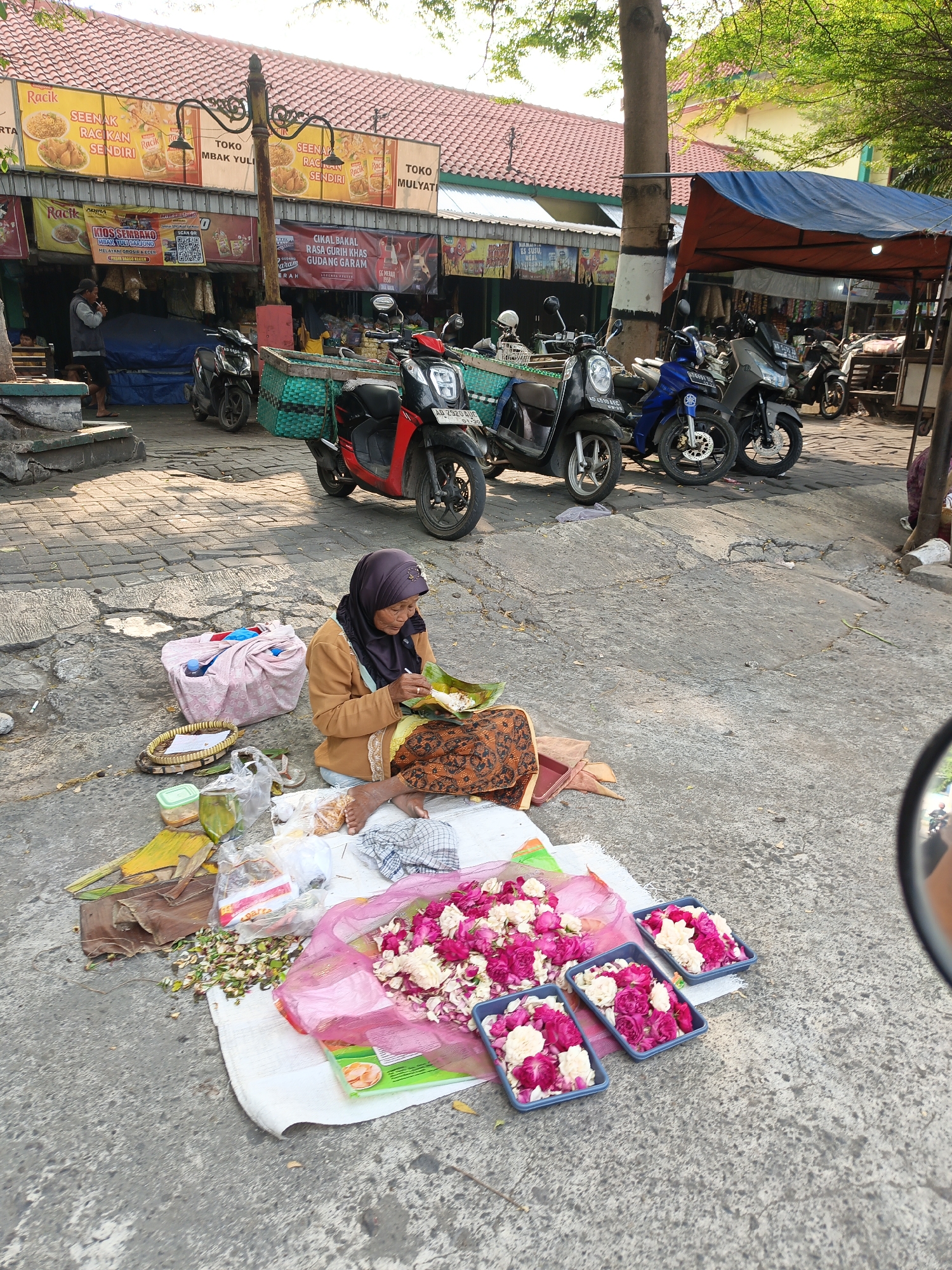 The image depicts a street scene in Klaten, Indonesia, likely a market area, with several shops visible in the background under a tiled roof. In the foreground, an elderly woman wearing a headscarf and a brown jacket sits on the ground, eating from a banana leaf parcel. Surrounding her are various items, including woven baskets, plastic bags containing food items, and piles of small flowers, predominantly pink and white, arranged in rectangular trays. Several motorcycles are parked on the paved street, with one red motorcycle prominently in the middle ground. The environment appears to be outdoors, with sunlight illuminating the scene and trees with green foliage visible overhead. The overall impression is that of a typical day in a local Indonesian marketplace.