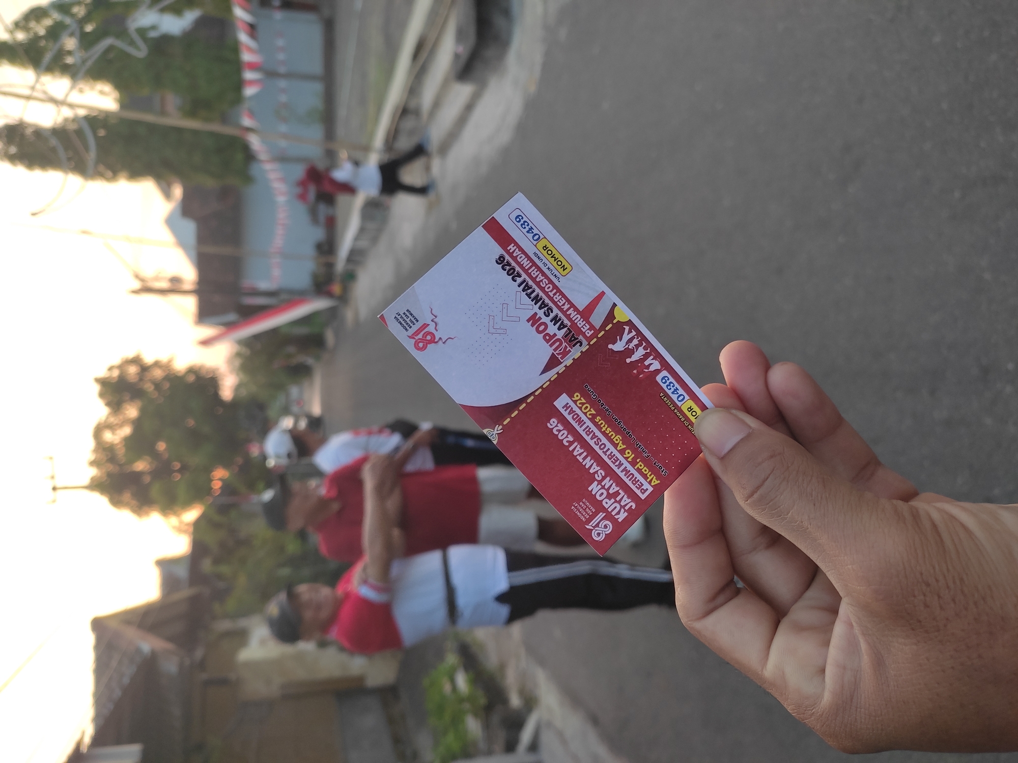 In the city of Cekok, Indonesia, at dawn, a hand holds up a red and white coupon. The coupon is for a "JALAN SANTAI" (leisure walk) event in 2026, named "PERUM KERTOSARI INDAH." The event is scheduled for Sunday, August 16th, 2026, with a start and finish line indicated. The coupon has a number "0439" visible. In the blurred background, several men in red and white shirts and caps stand on a street lined with houses. Indonesian flags are visible decorating the street, suggesting a national celebration or event. The overall ambiance is that of a community gathering or event being prepared for.