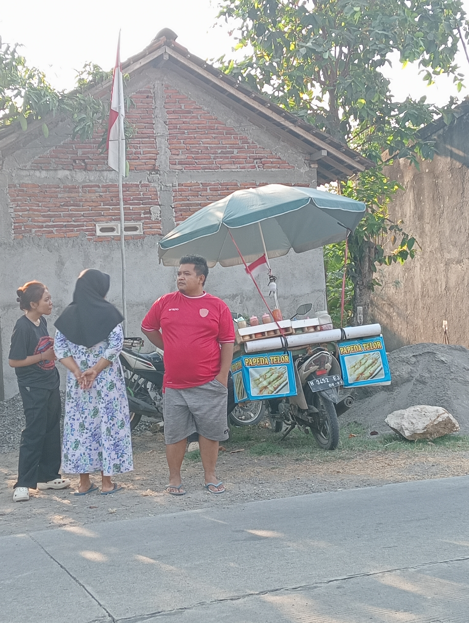 In Tanjunganyar, Indonesia, three individuals stand near a food cart attached to a motorcycle. The cart, shaded by a green umbrella, displays the text "PAPEDA TELOR" and features images of food. A man in a red shirt and gray shorts stands to the right, facing left with his hands in his pockets. To his left, a woman wearing a black hijab and a floral dress faces away from the camera. A third person, with their hair tied back and wearing a black t-shirt and black pants, stands to the far left, facing the other two. In the background, a brick building with a red-tiled roof is visible, with a flagpole displaying the Indonesian flag. The ground is a mixture of dirt and gravel, and a large rock rests to the right of the motorcycle. The scene is lit by the warm glow of what appears to be late afternoon sunlight.
