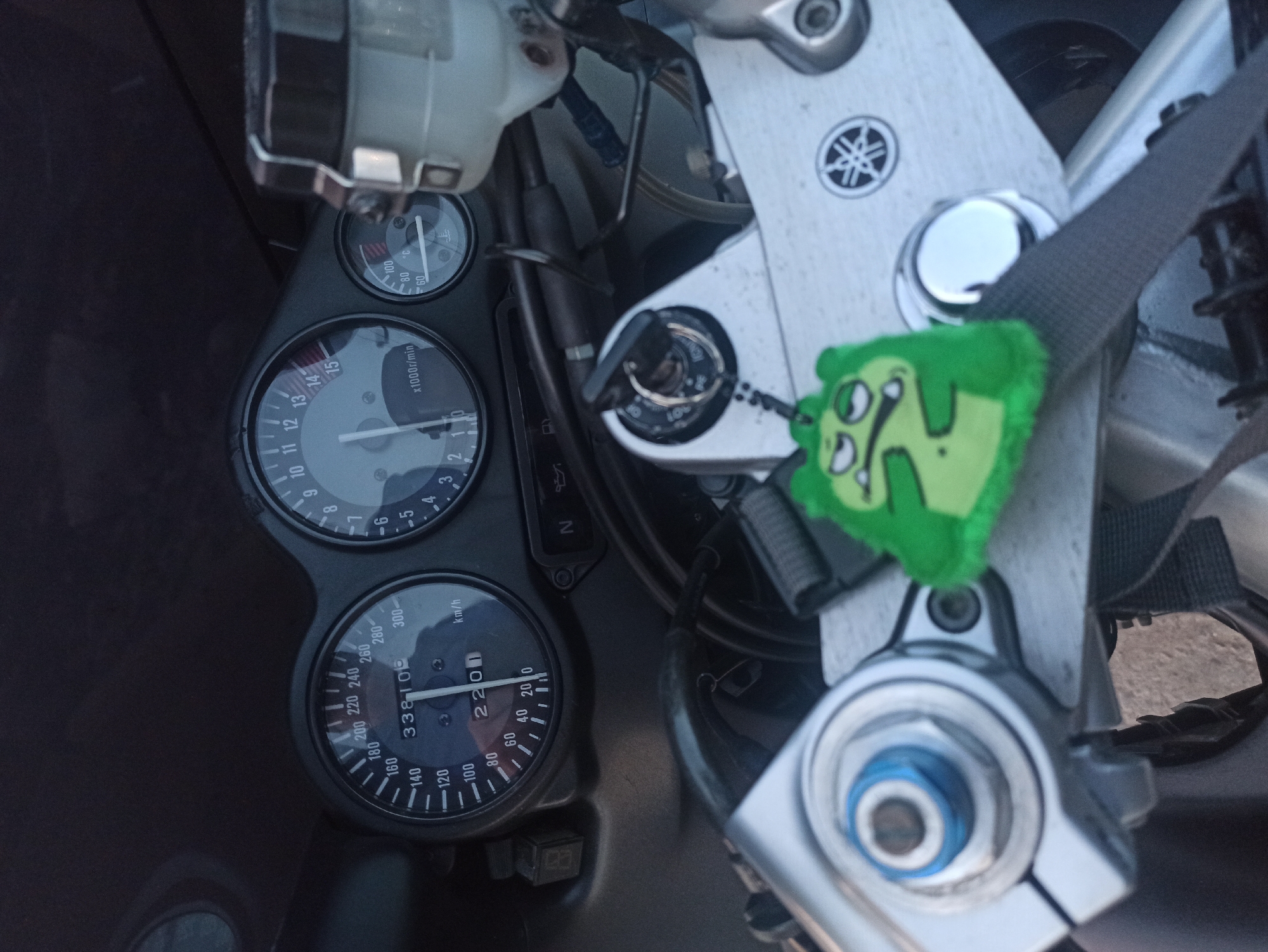 This is a close-up shot of a motorcycle's dashboard, indicating it is stationary. The dashboard features a speedometer with a reading of 2201 km/h and a tachometer showing about 1000 r/min. A key is inserted into the ignition switch, adorned with a green, cartoon-like keychain. A Yamaha logo is visible on the metal fixture above the gauges. The background is indistinct, with no clear indicators of the city or environment.