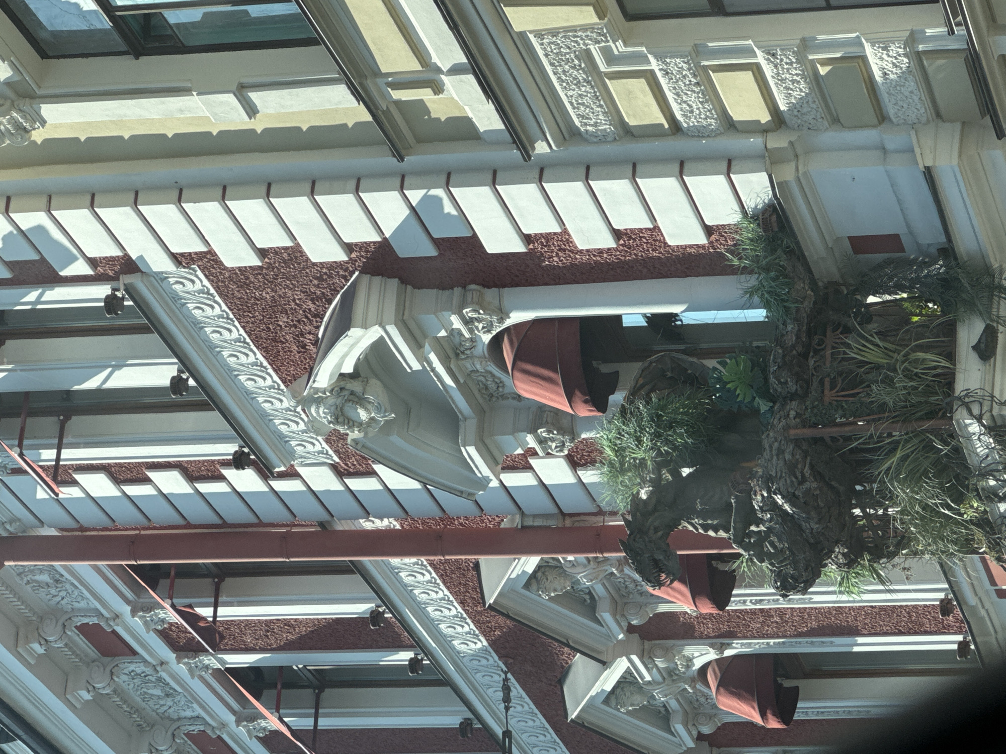 This media file presents an upward-angled view of a building's facade, likely in Saint Petersburg, Russia, given the context. The architecture features a textured maroon base beneath white, rectangular panels that alternate with darker, recessed sections, creating a sense of depth and pattern. Above this, decorative white moldings with intricate scrolling designs are visible, interspersed with what appear to be sculpted faces or figures.
Several red awnings are partially visible, adding a splash of color to the scene. Hanging plants, including lush greenery and what looks like a pine tree trunk, cascade down from an upper level, suggesting a green, possibly urban, setting. The sunlight indicates it is daytime, and the shadows suggest a sunny or partly cloudy day.
The style of the architecture suggests a historical or classical influence, common in European cities like Saint Petersburg. There is no discernible activity or event taking place; the scene appears to be a static architectural study. No text is visible in the image. The overall impression is one of ornate and well-maintained urban architecture.