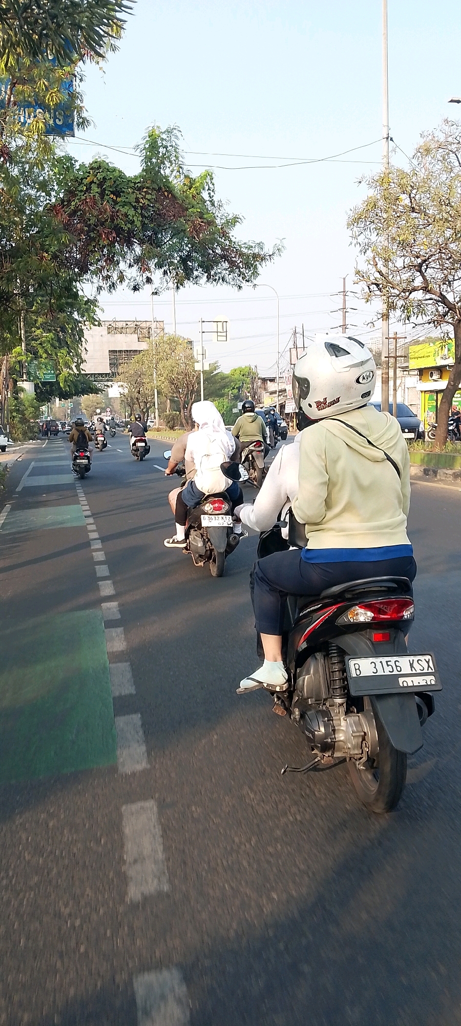 In Bekasi, Indonesia, a group of individuals are riding motorbikes on a paved road during the daytime. The perspective is from a motorbike rider, looking forward and slightly to the right. In the foreground, a rider wearing a light yellow hoodie and dark pants is on a black scooter, with a license plate visible that reads "B 3156 KSX". To the left and slightly ahead, another rider in a gray shirt and white hijab is on a scooter. Behind them, several other motorbikes are visible, carrying one or two passengers each. Trees line the sides of the road, and buildings and power lines can be seen in the background. The sky is clear and bright, suggesting good weather. The road is marked with white dashed lines and a green lane on the left side.