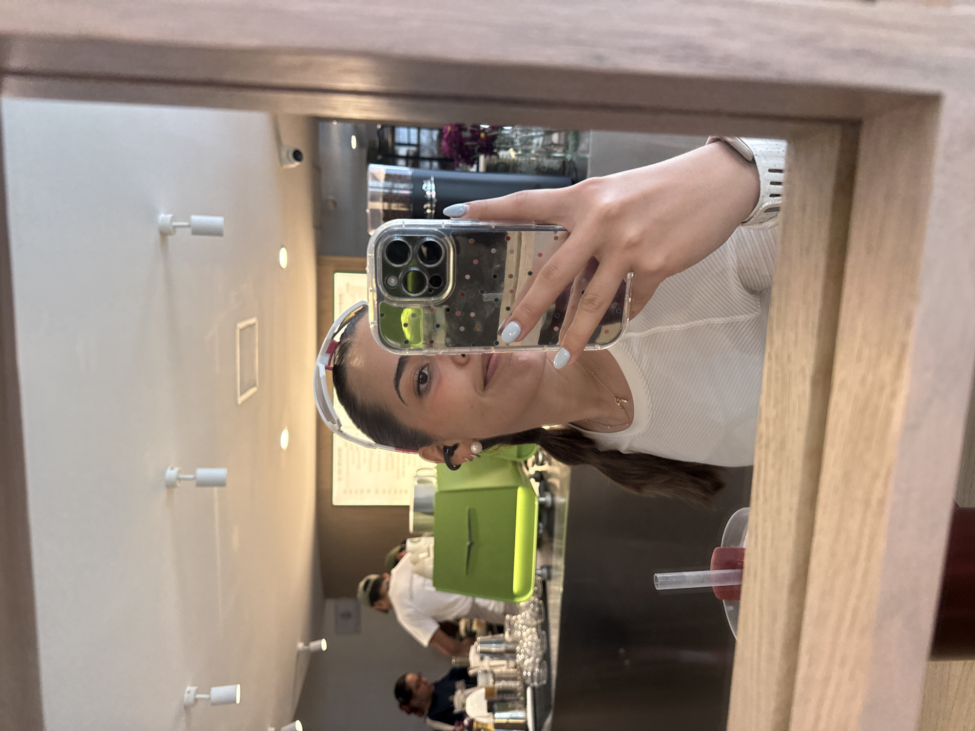 A person with tan skin is holding an iPhone in a clear polka-dot case, taking a selfie in a mirror. The person is wearing a white ribbed shirt and a white and red visor. Their hair is pulled back into a ponytail. They are wearing a silver watch on their left wrist. The background shows what appears to be the interior of a cafe or juice bar. There is a line of clear glasses on the counter in front of a green juice press. Two people are visible behind the counter, both wearing white shirts. The walls are white with track lighting. Through the reflection, various objects and elements are seen which might identify the place as Puebla, Mexico. There is a cup with a straw placed in the foreground of the mirror.