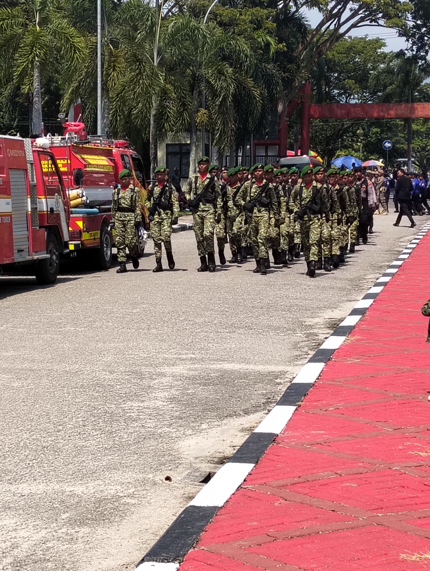 In Parigi, Indonesia, a contingent of approximately 30 uniformed individuals, likely military personnel based on their camouflage uniforms and green berets, march in formation on a paved road. They are armed with rifles. To their left, a red fire truck is parked. The scene appears to be part of a parade or official event, as suggested by the decorated red and black curbs bordering the walkway on the right. Palm trees and other foliage form a backdrop, indicating a tropical environment. The sky is bright and appears to be daytime.