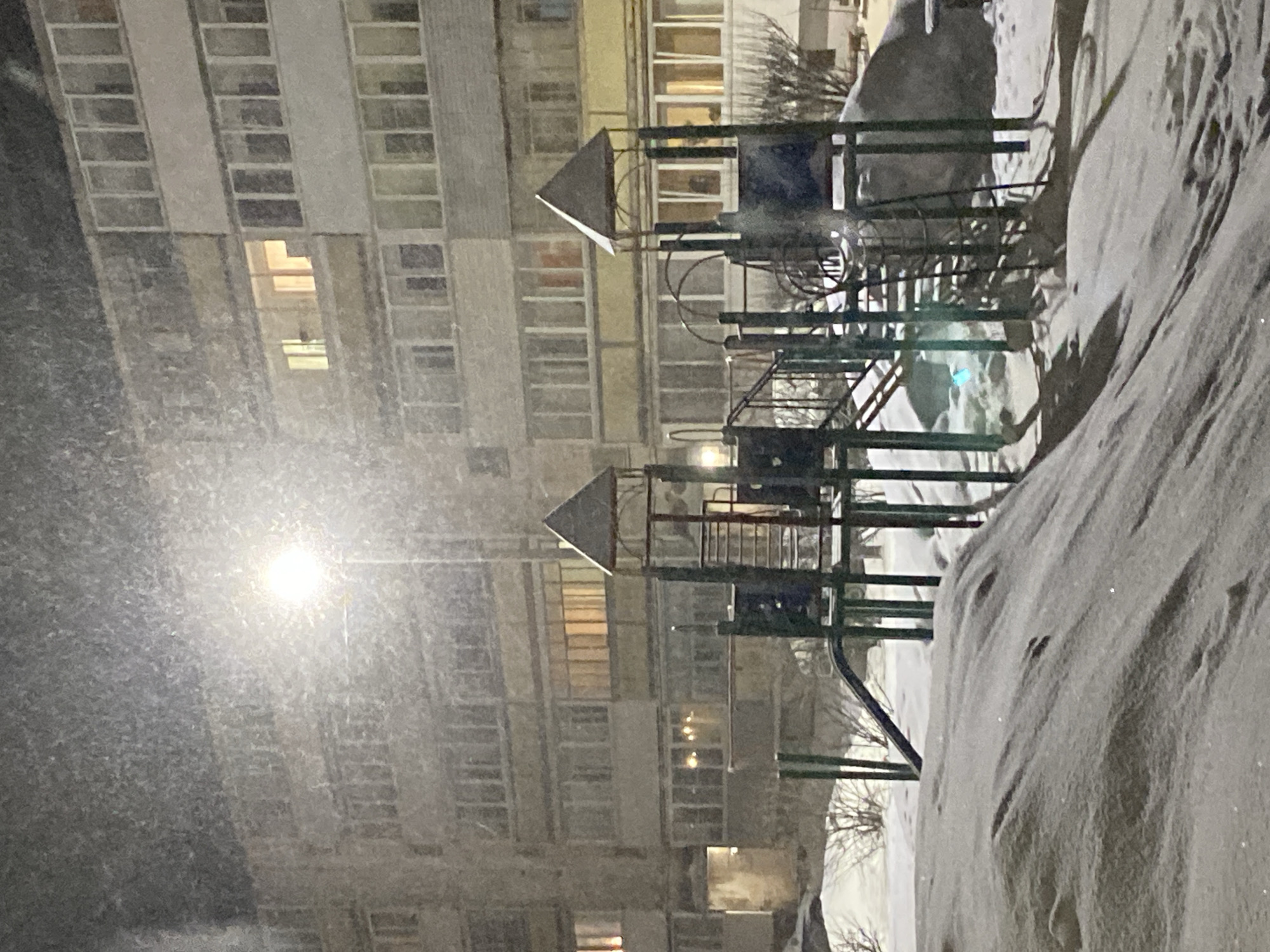 The image depicts a snow-covered playground in Noviy Urengoy, Russia, at night. The playground is situated near a multi-story apartment building with illuminated windows. A streetlight shines brightly, partially obscured by falling snow. The playground equipment is covered in snowdrifts. The scene is quiet and still.